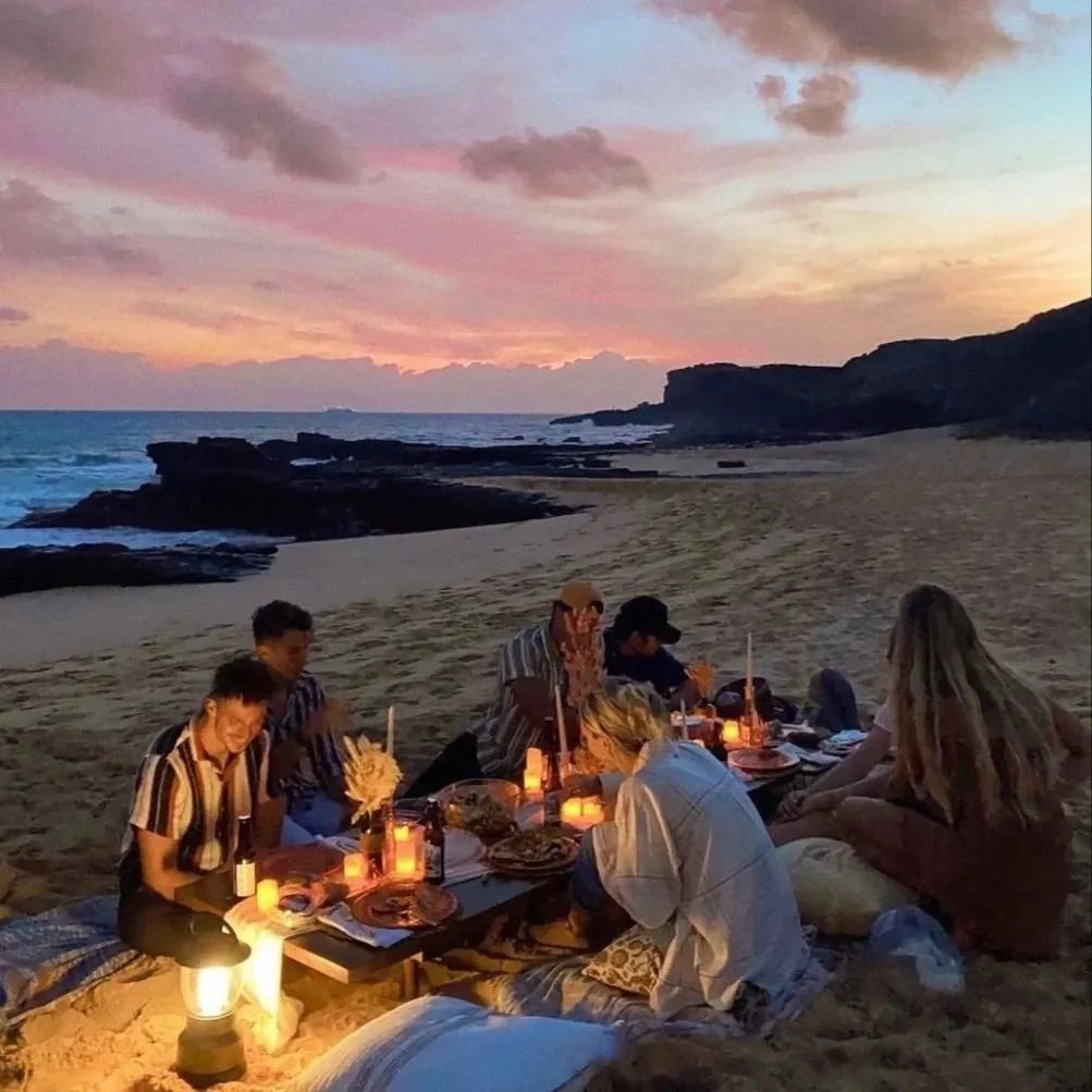 A group of people sitting on the sand at a beach during sunset, enjoying a meal by candlelight with the ocean and cliffs in the background.
