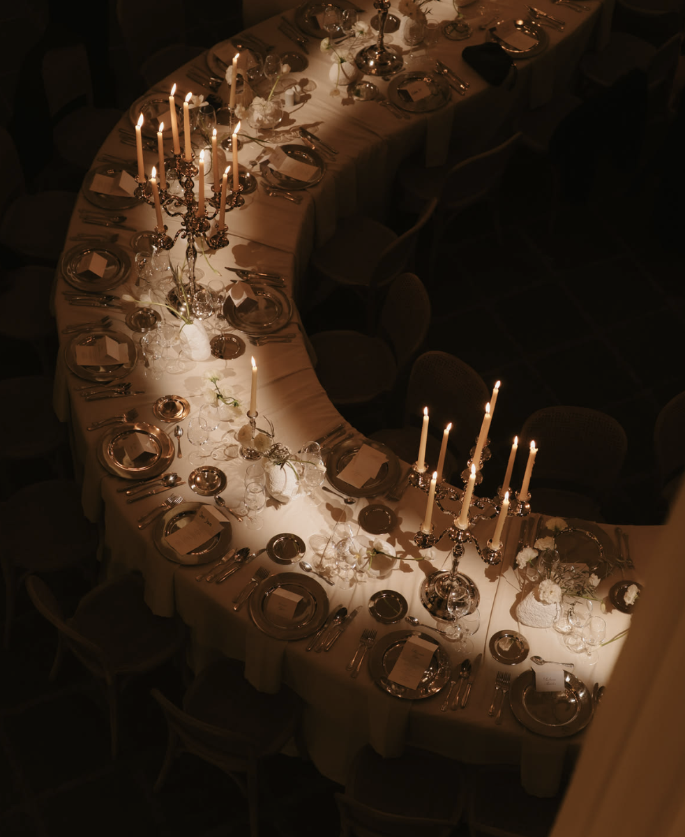 Overhead view of a long, semi-circular dining table set for a formal dinner with lit candelabras, plates, silverware, glasses, and decorative centerpieces in a dimly lit room.