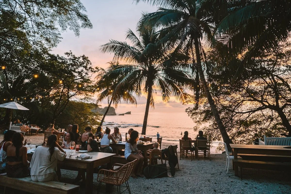 People dining at a beachside restaurant during sunset, with palm trees and ocean in the background.