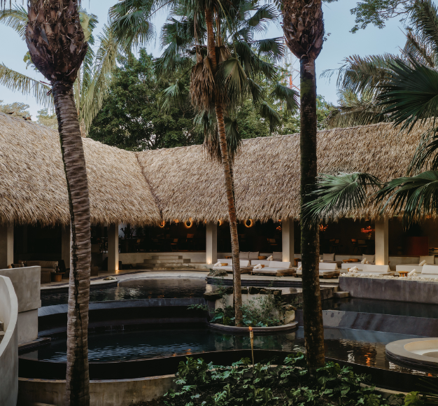 A luxurious outdoor pool area with thatched-roof cabanas, surrounded by palm trees and lush greenery, creating a tropical resort vibe.