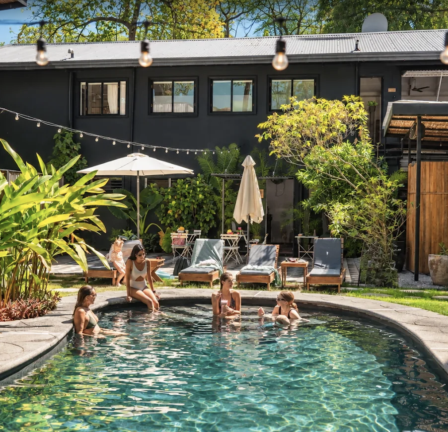 People relaxing and swimming in a backyard swimming pool, with outdoor lounge chairs, umbrellas, and lush greenery in the background.
