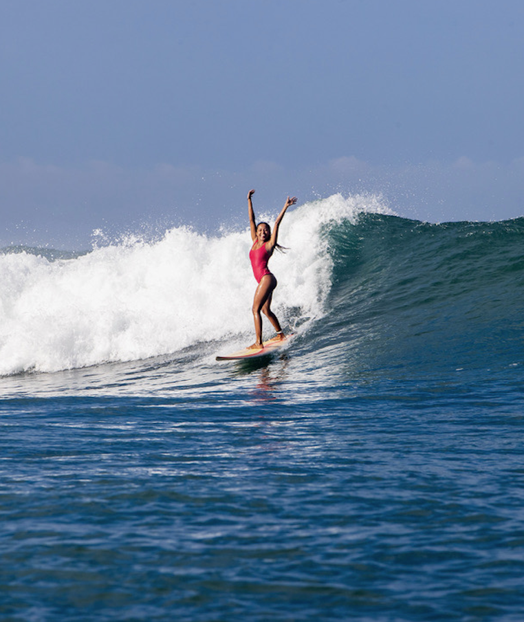 A woman in a pink swimsuit surfing on a wave in the ocean under a clear blue sky.