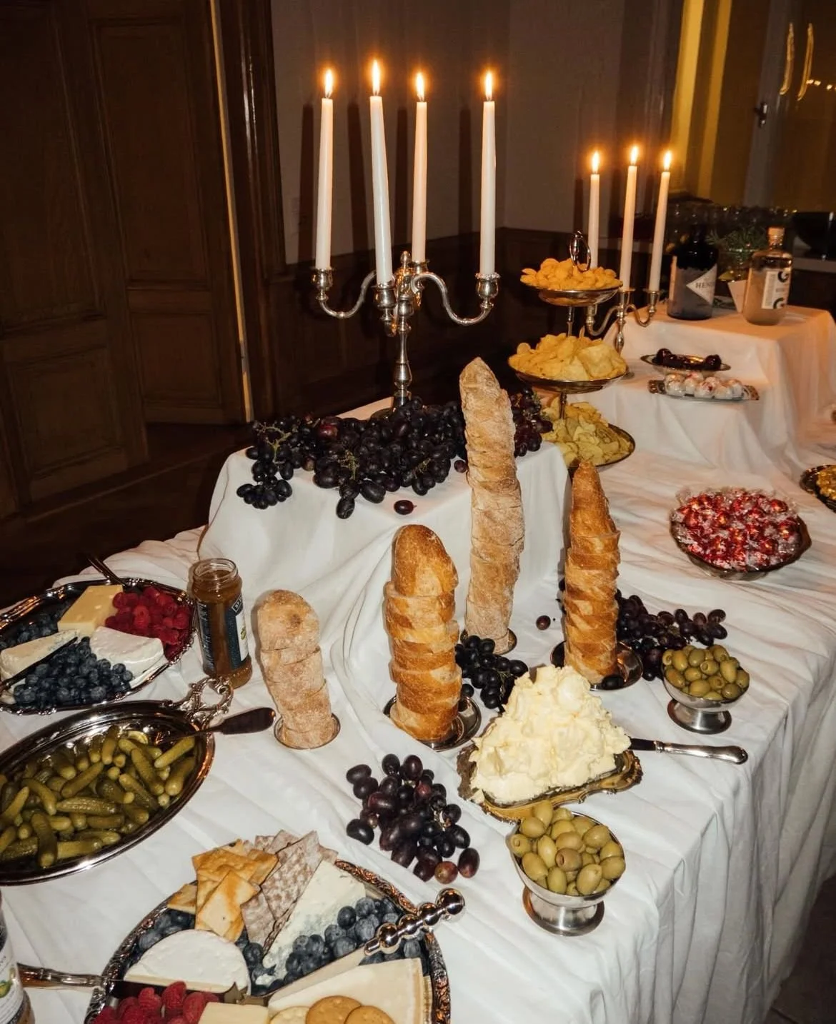 An elaborate spread of food on a table decorated with a white tablecloth, featuring bread, cheeses, grapes, olives, chips, and a large candelabrum with lit candles.