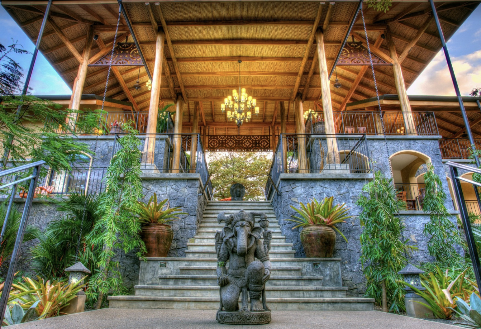 View of a multi-level outdoor pavilion with stone steps leading up to a wooden deck with a chandelier, greenery, and decorative elements, including a carved elephant statue at the bottom of the stairs.
