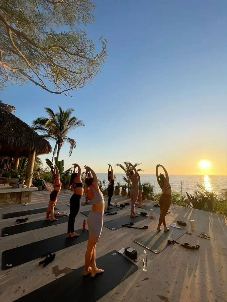 Group of women practicing yoga outdoors on mats by the ocean at sunset, surrounded by palm trees and tropical plants.