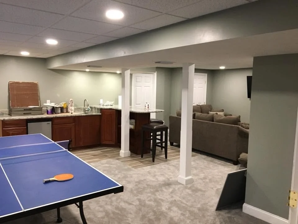 A basement room with a ping pong table and a ping pong paddle on it, a kitchenette with a sink, microwave, and countertop, a small seating area with sofas, and a television on the wall.