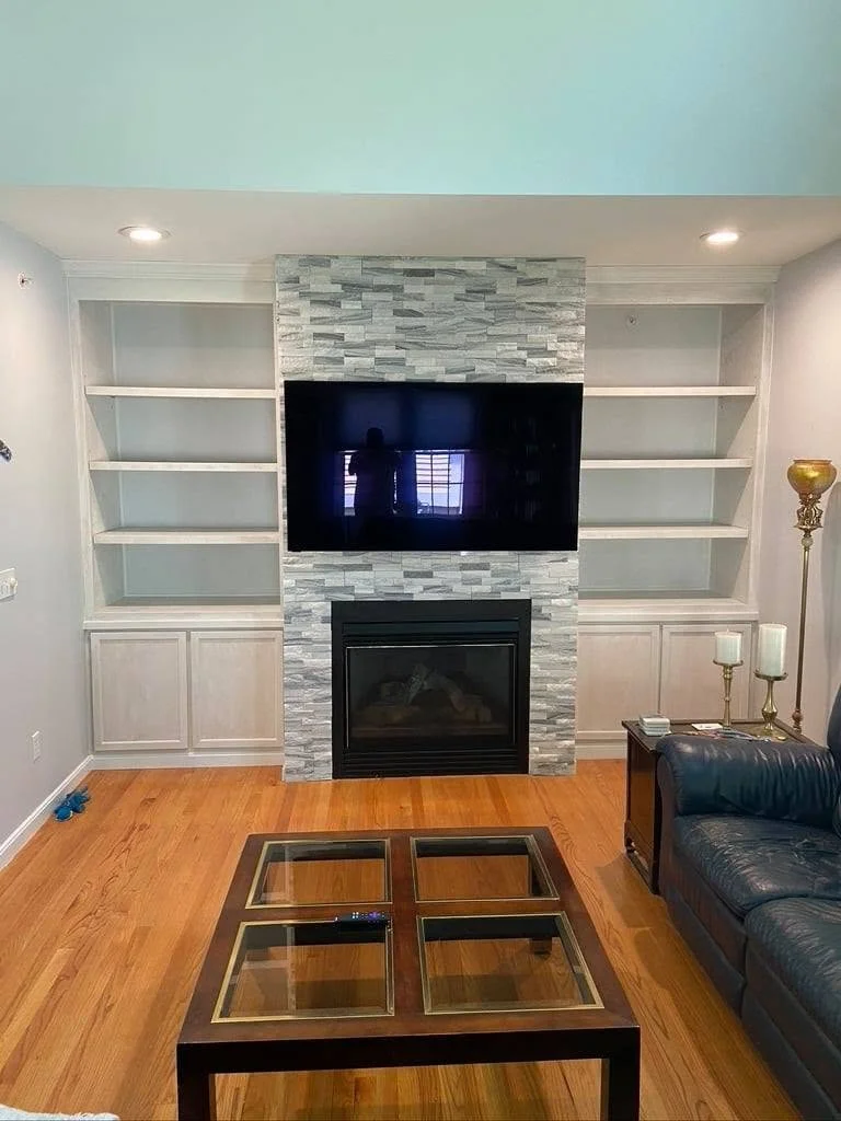 Living room with a stone fireplace, TV mounted above, and empty white bookshelves on each side. There is a leather sofa, glass-top coffee table, and a brass floor lamp with candles on a side table.