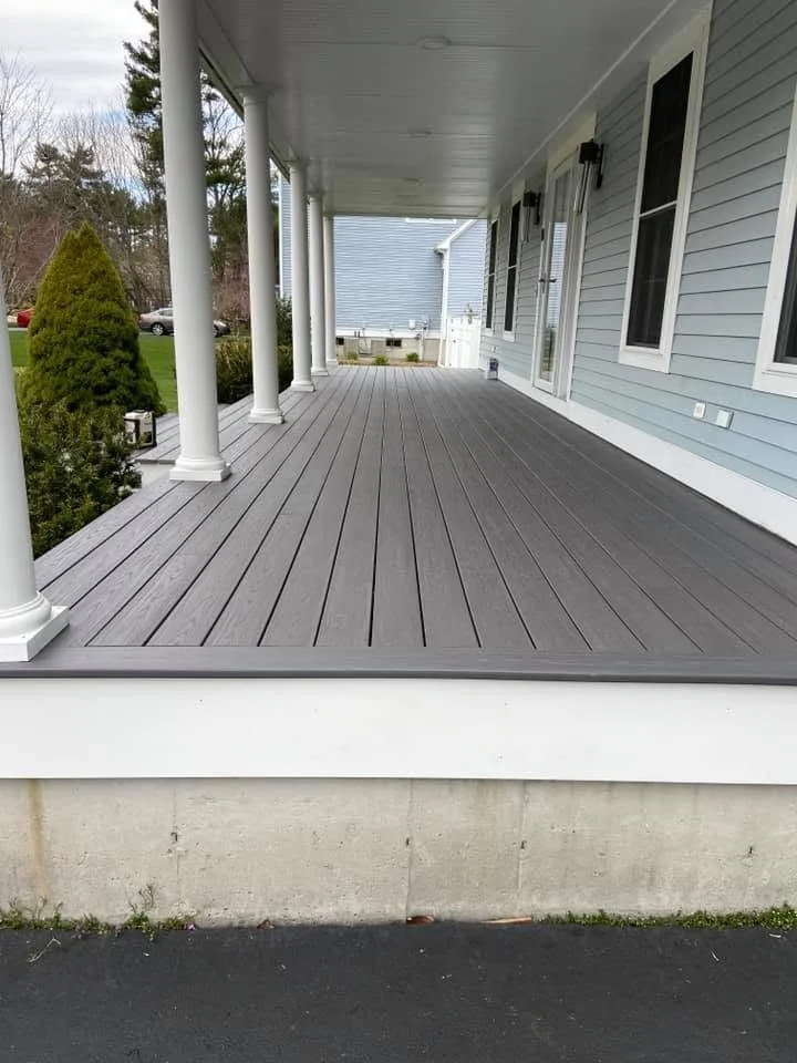 A spacious front porch with gray wooden flooring, white support columns, and light blue siding house exterior with black outdoor wall lanterns.