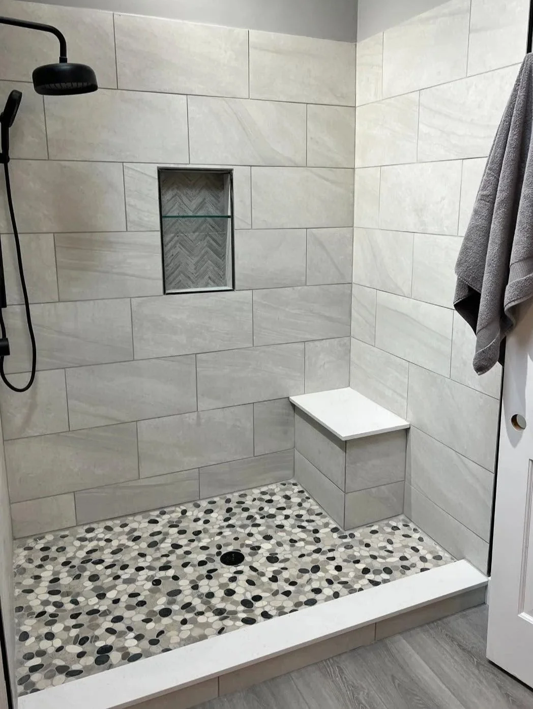 A modern walk-in shower with large beige tiles, a built-in niche with herringbone patterned tile, pebble tile floor, a black rainfall showerhead, and a white bench with a towel hanging on a hook outside.