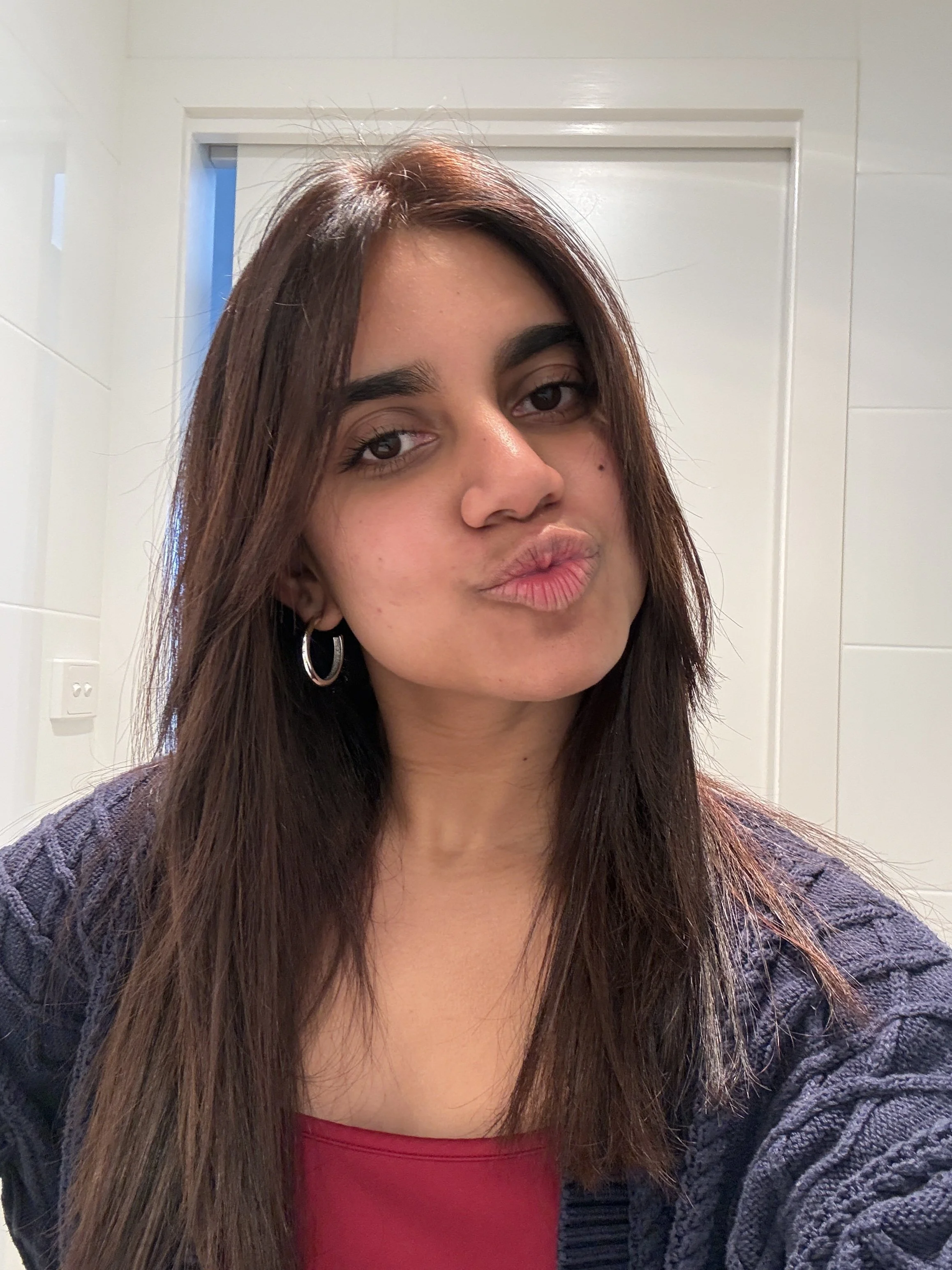 Close-up selfie of a woman with long brown hair making a pouting expression, wearing hoop earrings and a dark cardigan over a red top.