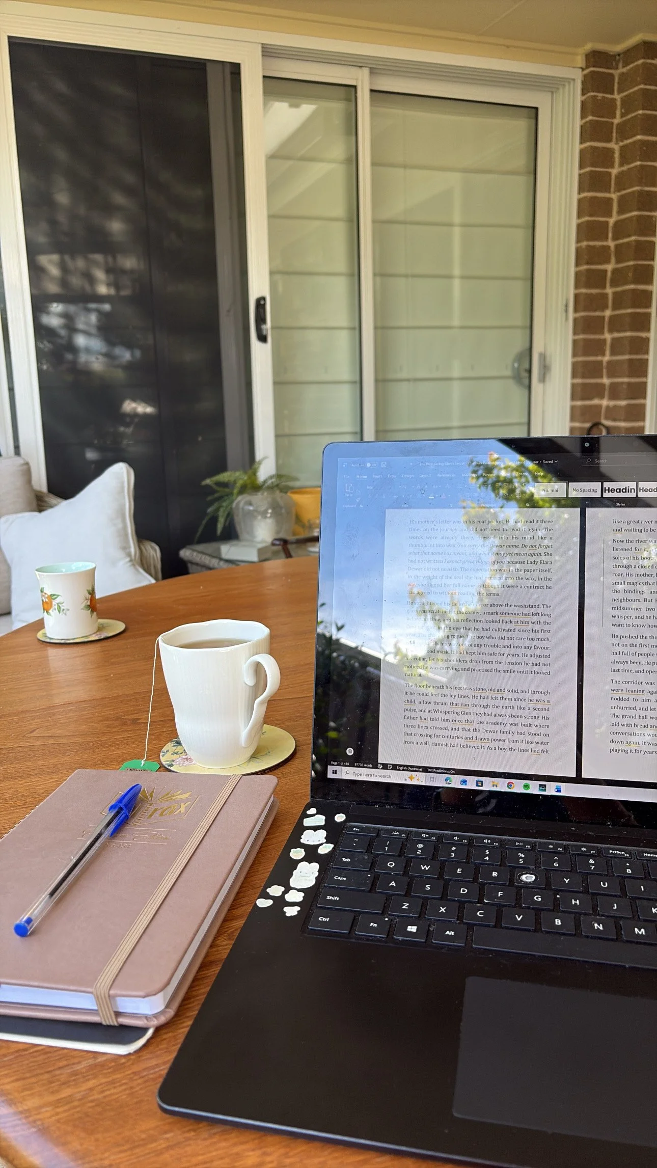 Laptop displaying a document on a wooden table with two cups of tea, a notebook, and a pen beside a window.