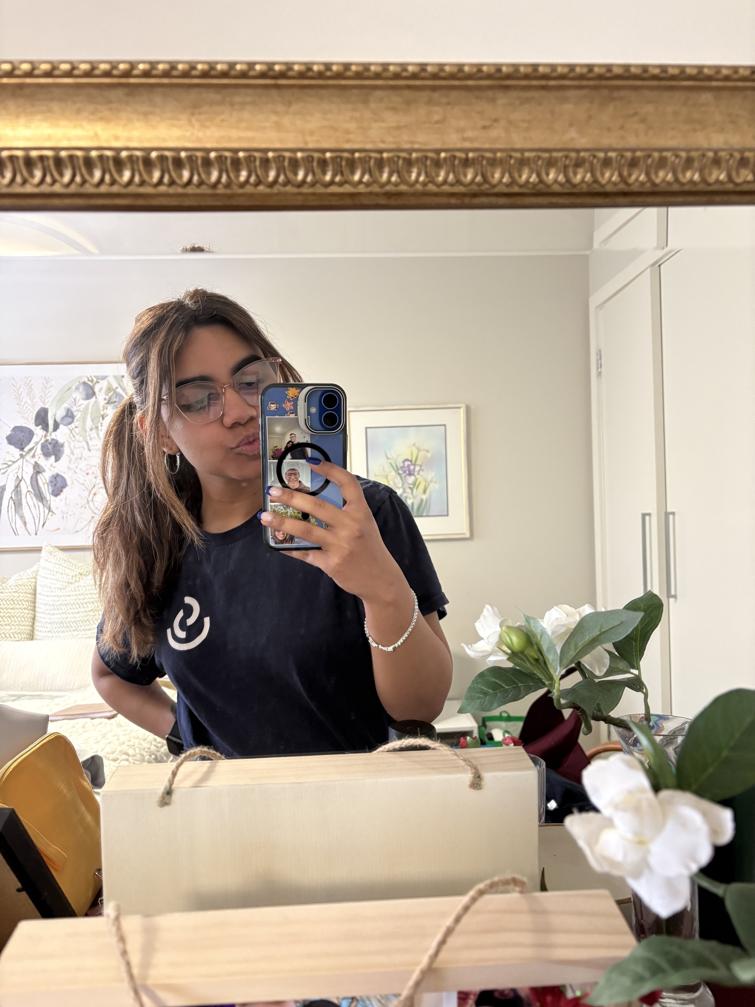 Person wearing glasses takes a mirror selfie with a decorated smartphone in a bright room with framed artwork, flowers, and wooden gift boxes.