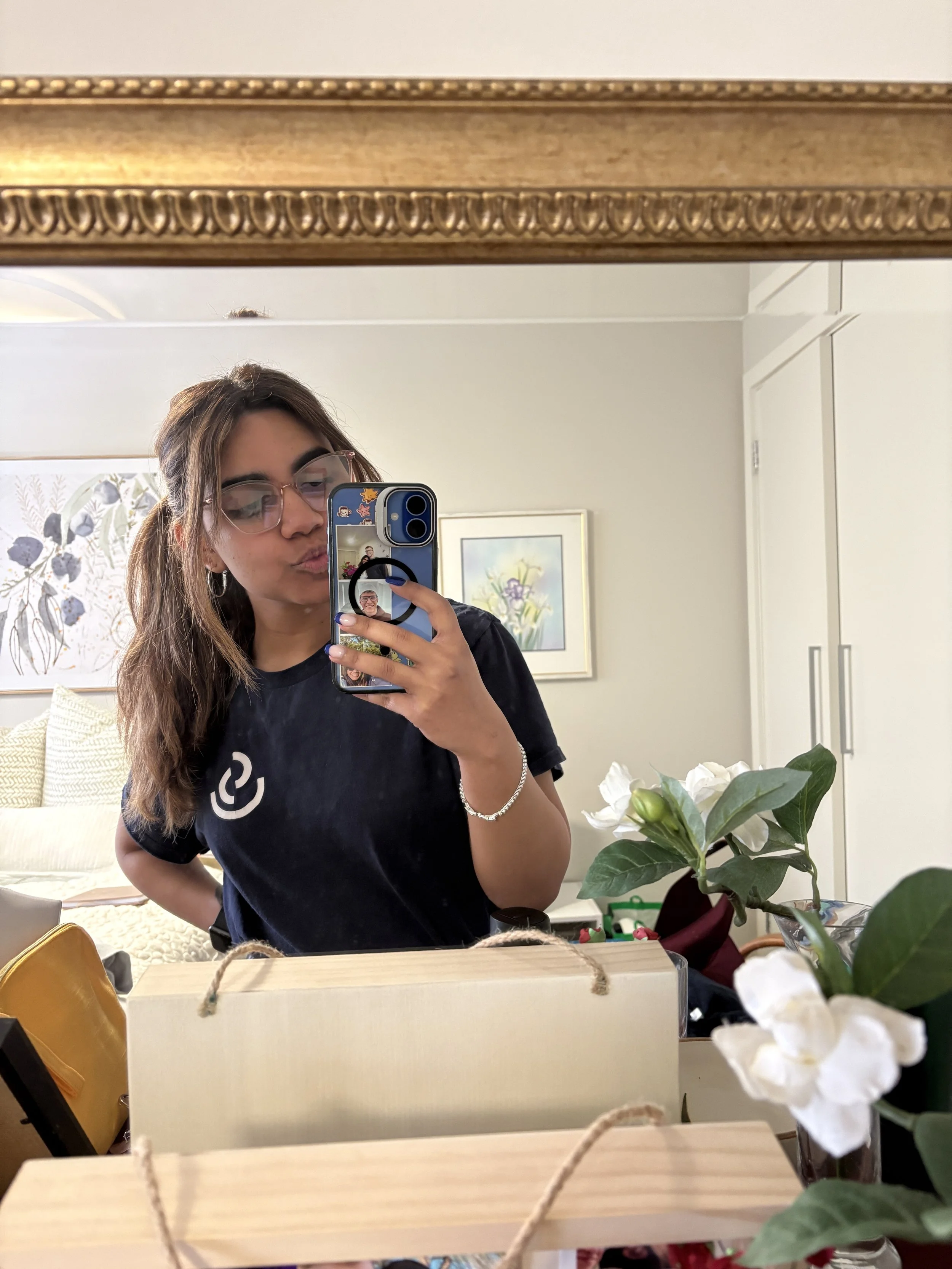 Person wearing glasses takes a mirror selfie with a smartphone in a bright room, with framed artwork, flowers, and gift boxes visible.