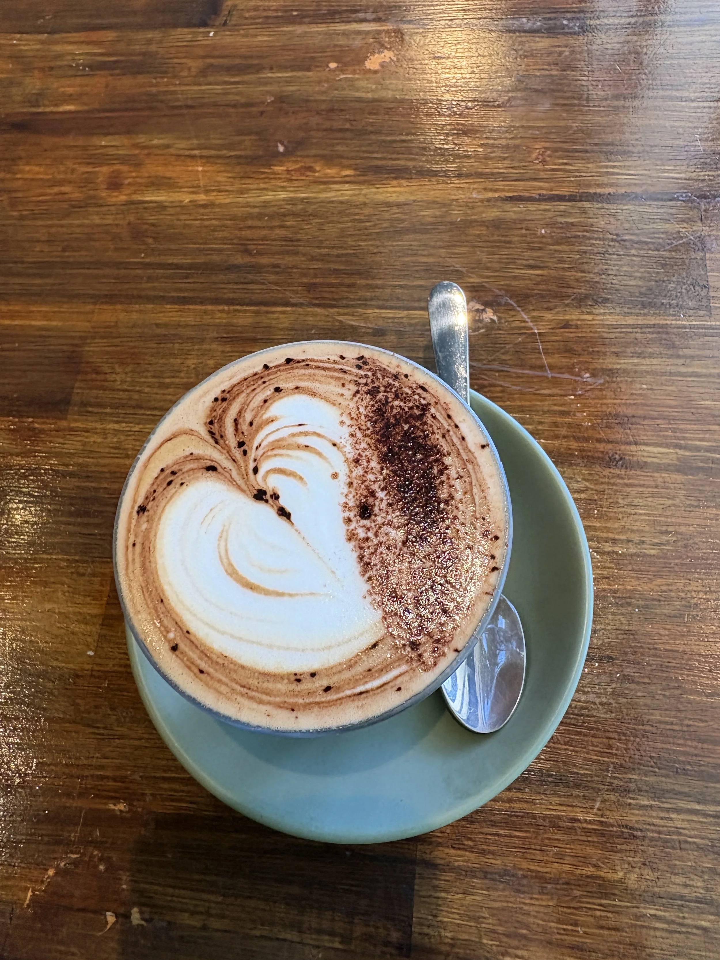 Top-down view of a chocolate cappuccino with foam art and cocoa powder on a blue saucer, alongside a spoon on a wooden table.