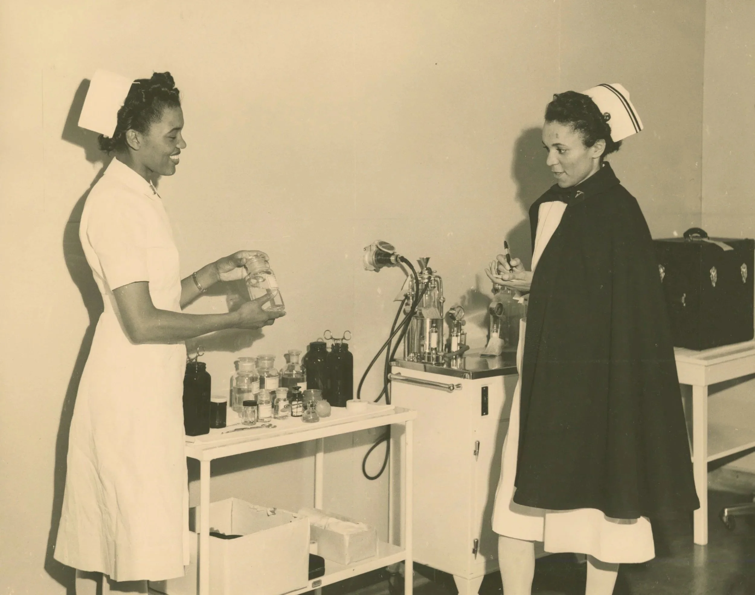 Black and white photo of two women wearing uniforms, possibly nurses, exchanging items in a medical or laboratory setting with bottles and equipment on a table.