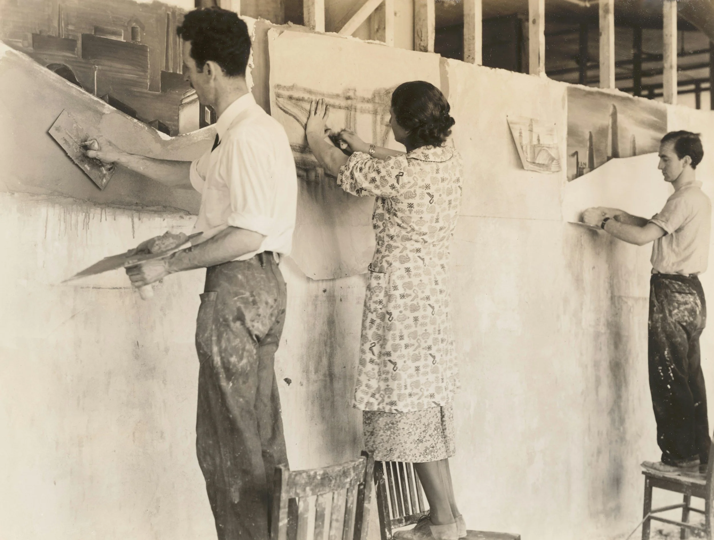 Three women and one man working on a large stone or sculpture in a workshop or art studio. The women are drawing or carving on the stone, while the man appears to be carving or chiseling the surface.