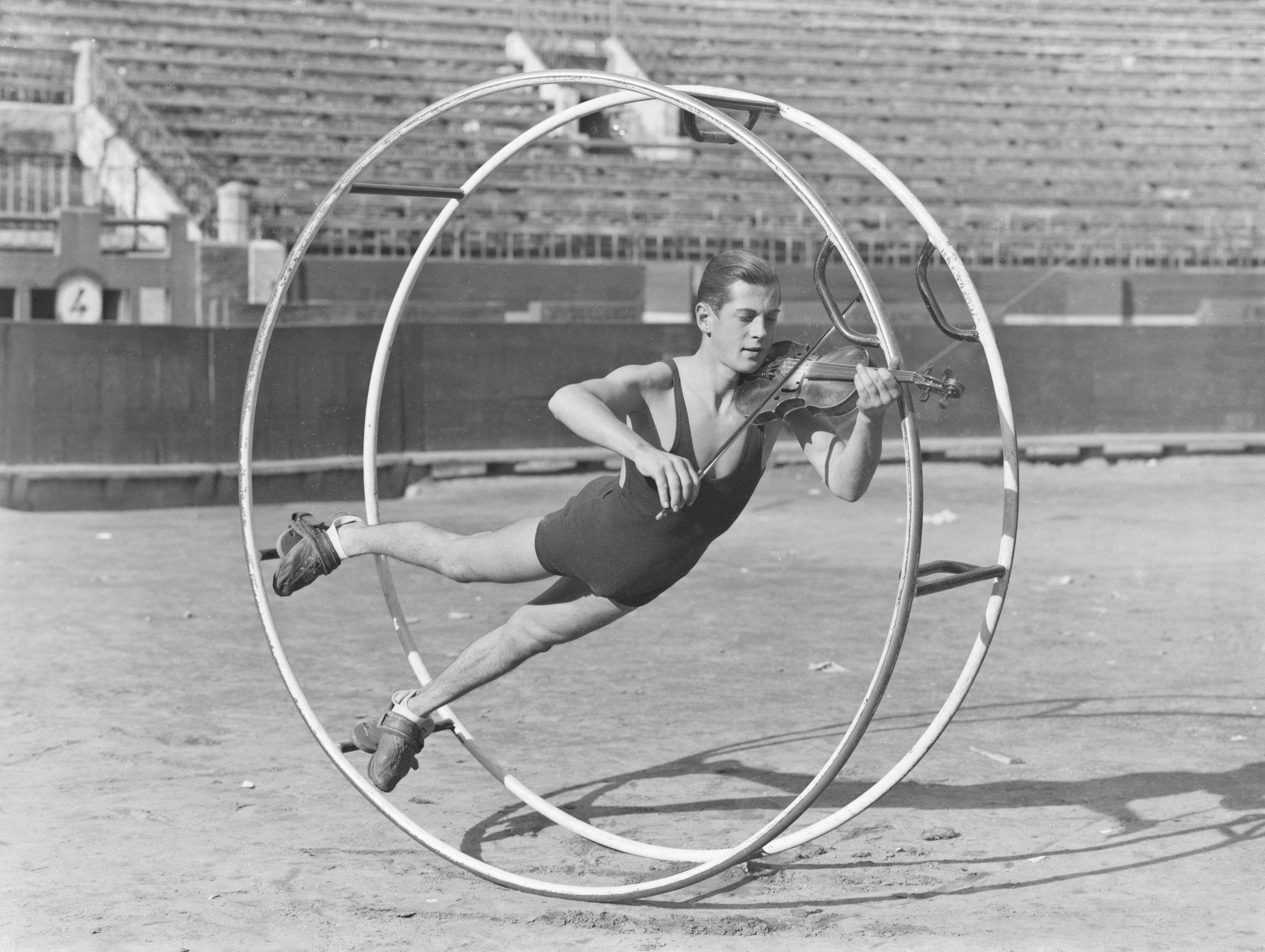 A woman in athletic clothing is performing a balancing exercise while lying inside a large metal wheel on an outdoor track. She is holding a violin and bow while balancing on her stomach.
