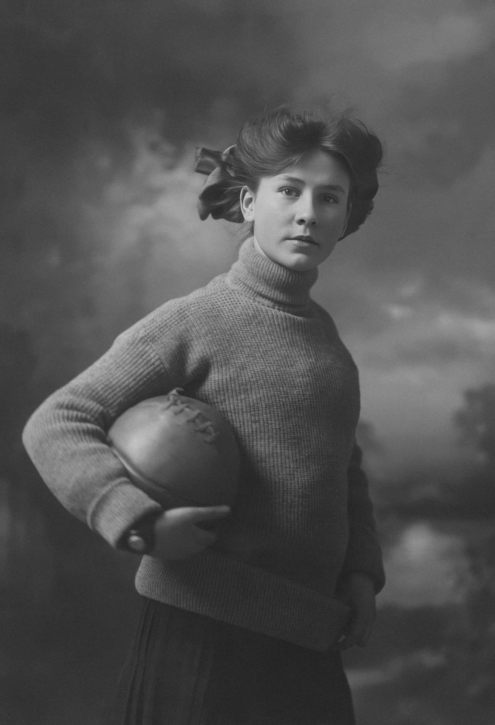 A black and white photograph of a woman wearing a turtleneck sweater and holding a football under her arm. Her hair is swept back, and she has a calm expression, standing against a cloudy sky background.