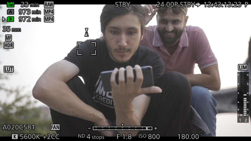 Two young men looking at a smartphone outdoors during daylight, one sitting and the other leaning over, both smiling and interested in the screen.
