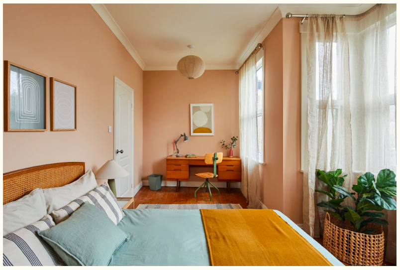 A cozy bedroom with a wooden bed, light blue bedding, and striped pillows. There is a white bedside lamp, framed artwork on the peach-colored wall, and a wooden desk with a green chair near large windows with sheer curtains. A large potted plant is in a woven basket, and a hanging paper lantern is on the ceiling.