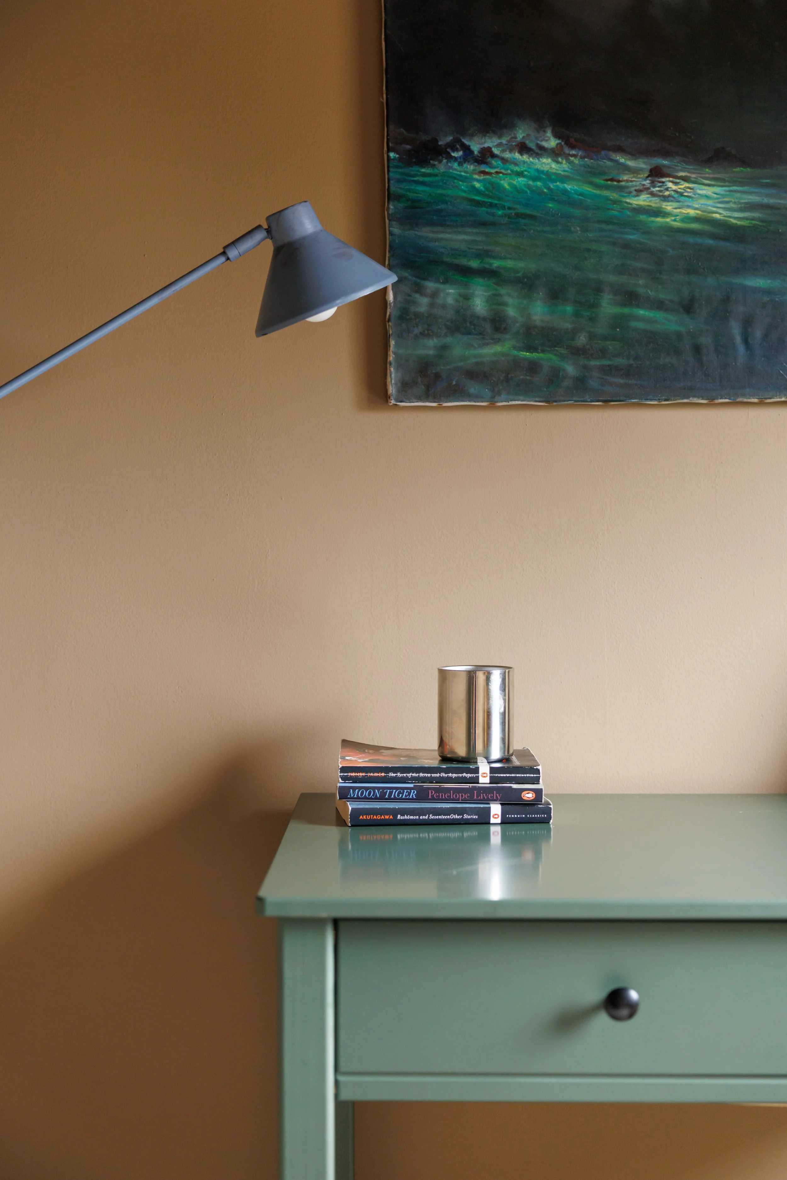 A mint green side table with a drawer, on which a stack of books is placed, topped with a silver canister. Above the table, a beige wall with a painting of a turbulent sea and stormy sky is visible, along with a silver desk lamp directed downward.