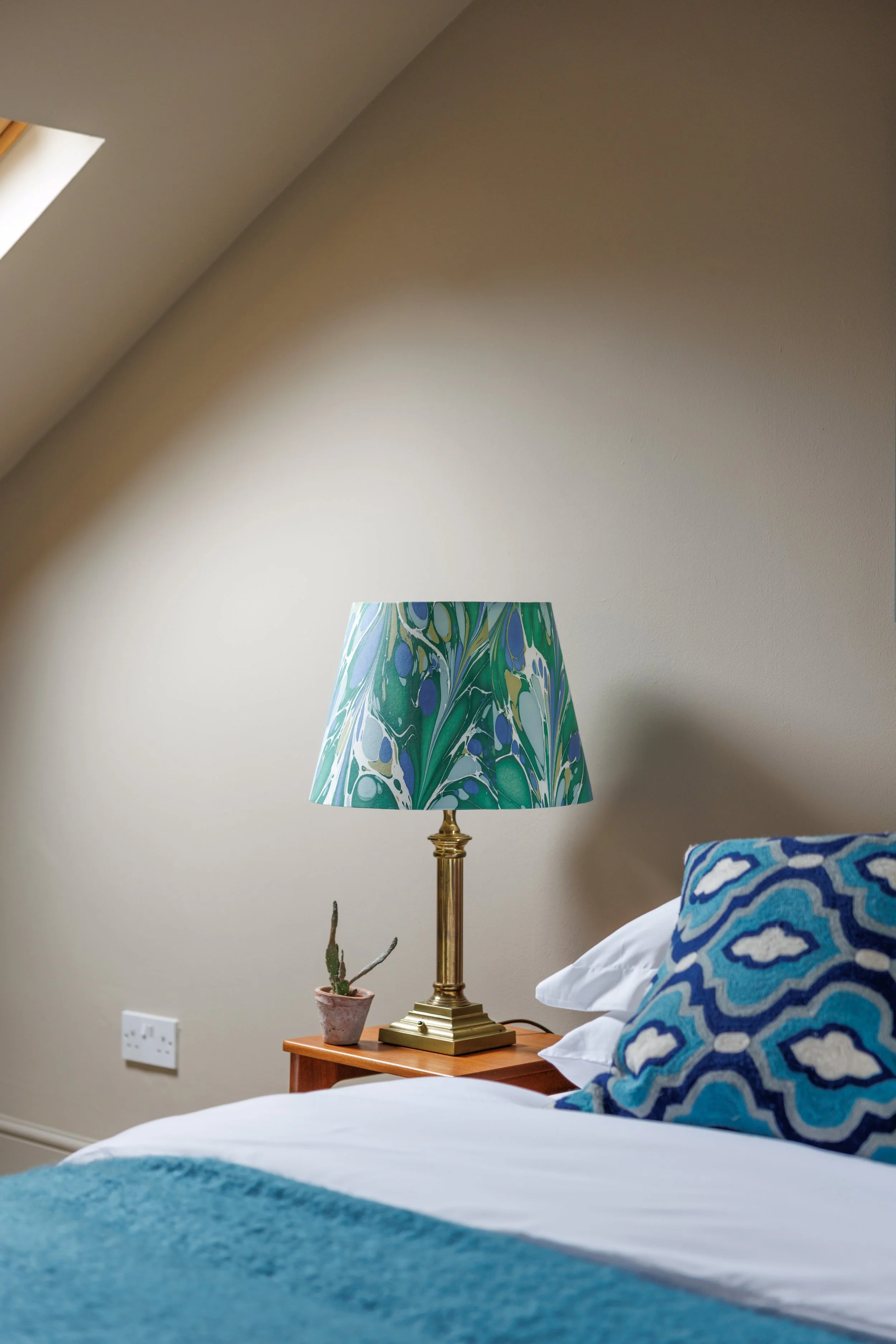 A bedroom corner with a bed and blue patterned pillows, wooden nightstand with a potted cactus, a gold lamp with a multicolored lampshade, and a beige wall with a skylight window.