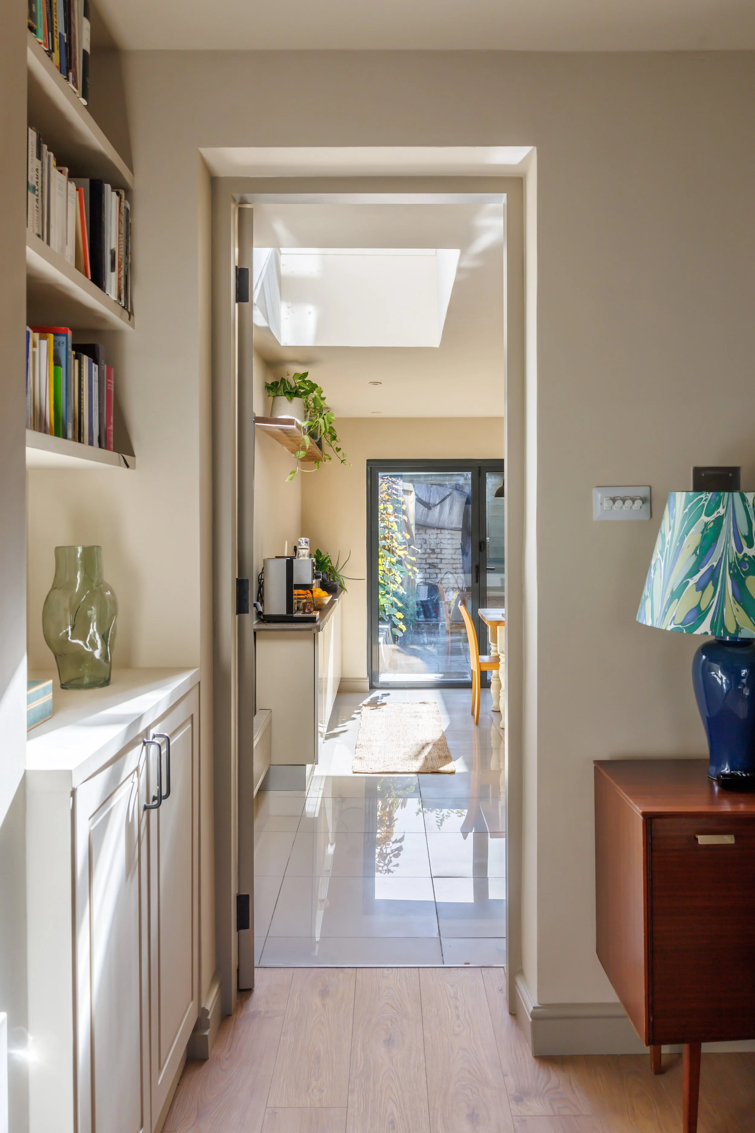 A bright corridor with a white tiled floor and beige walls, leading to a glass sliding door with a view of an outdoor area, decorated with indoor plants and hanging shelves with books and kitchen appliances.