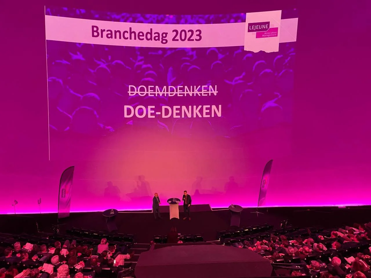 Podium tijdens Branchendag 2023 met de tekst ‘Doe-denken’, twee sprekers en een publiek in de zaal.