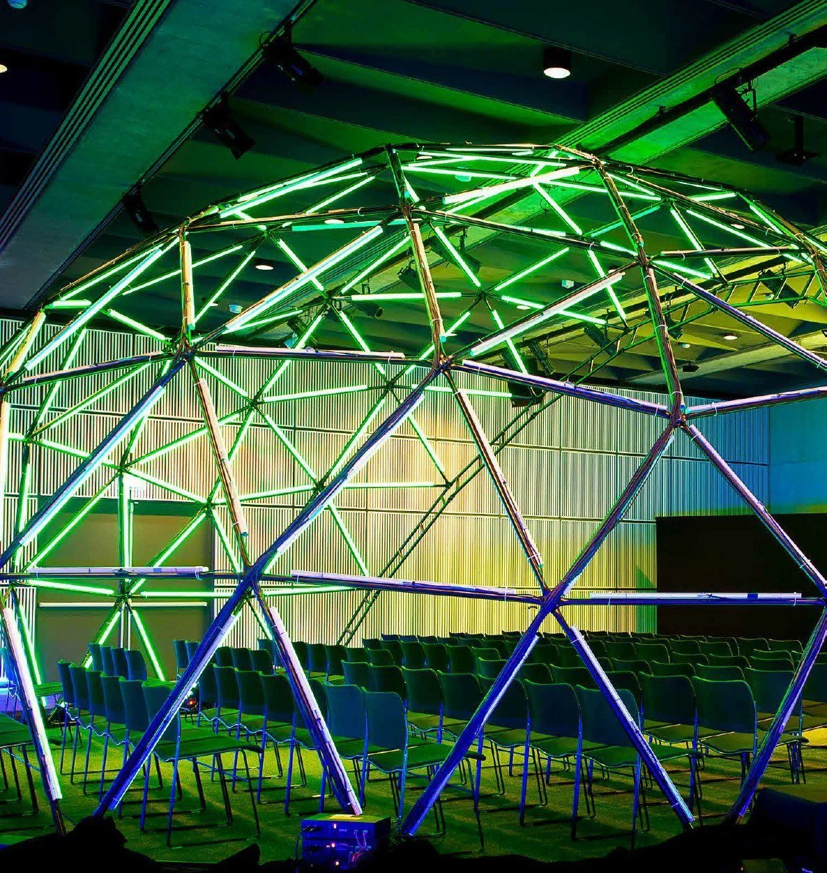 Een futuristische geometrische lichtinstallatie met groene neonbuizen boven rijen stoelen in een evenementenruimte.