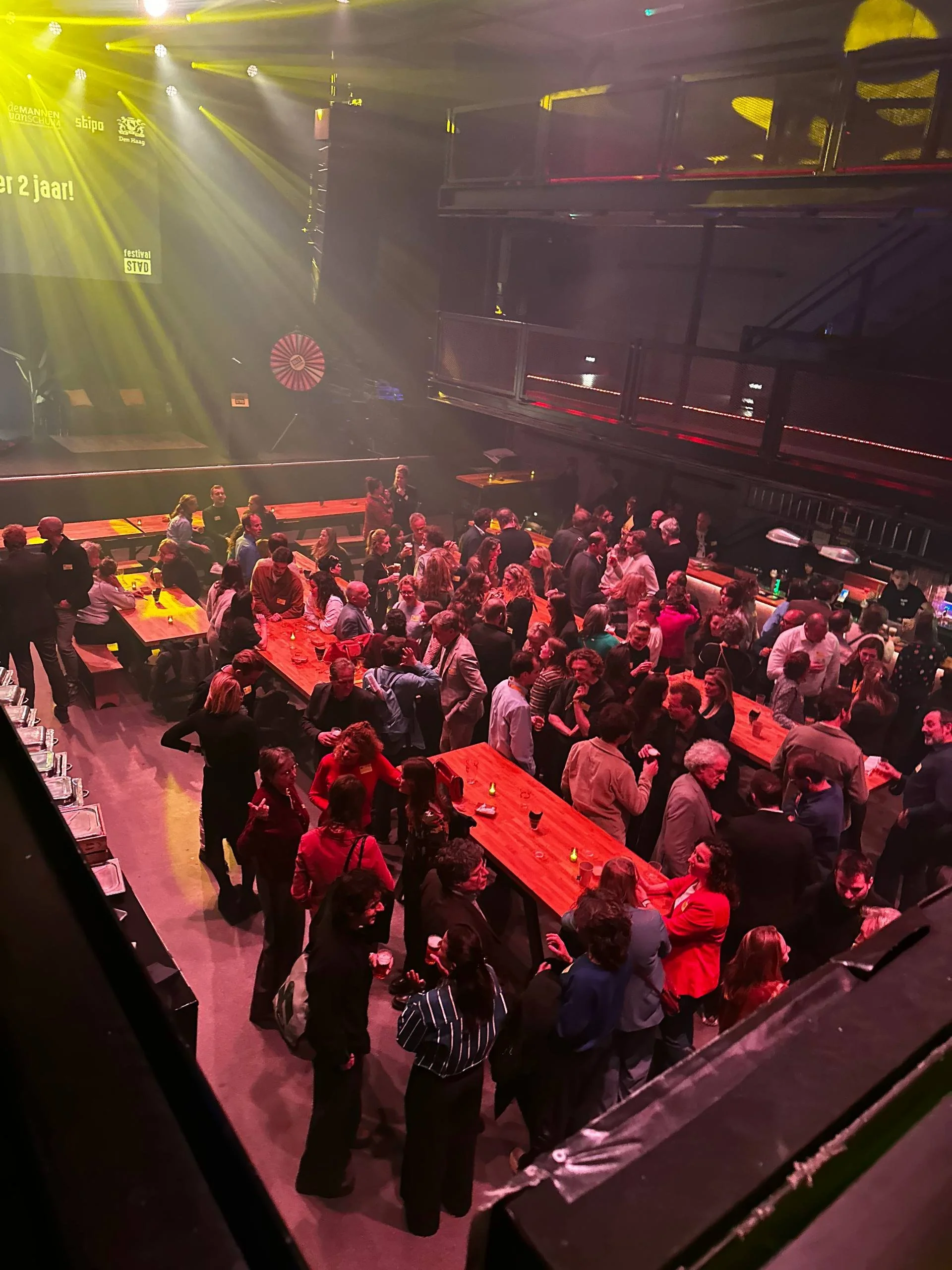 Drukbezocht evenement in een donkere zaal met lange tafels, pratende bezoekers en gele podiumverlichting.