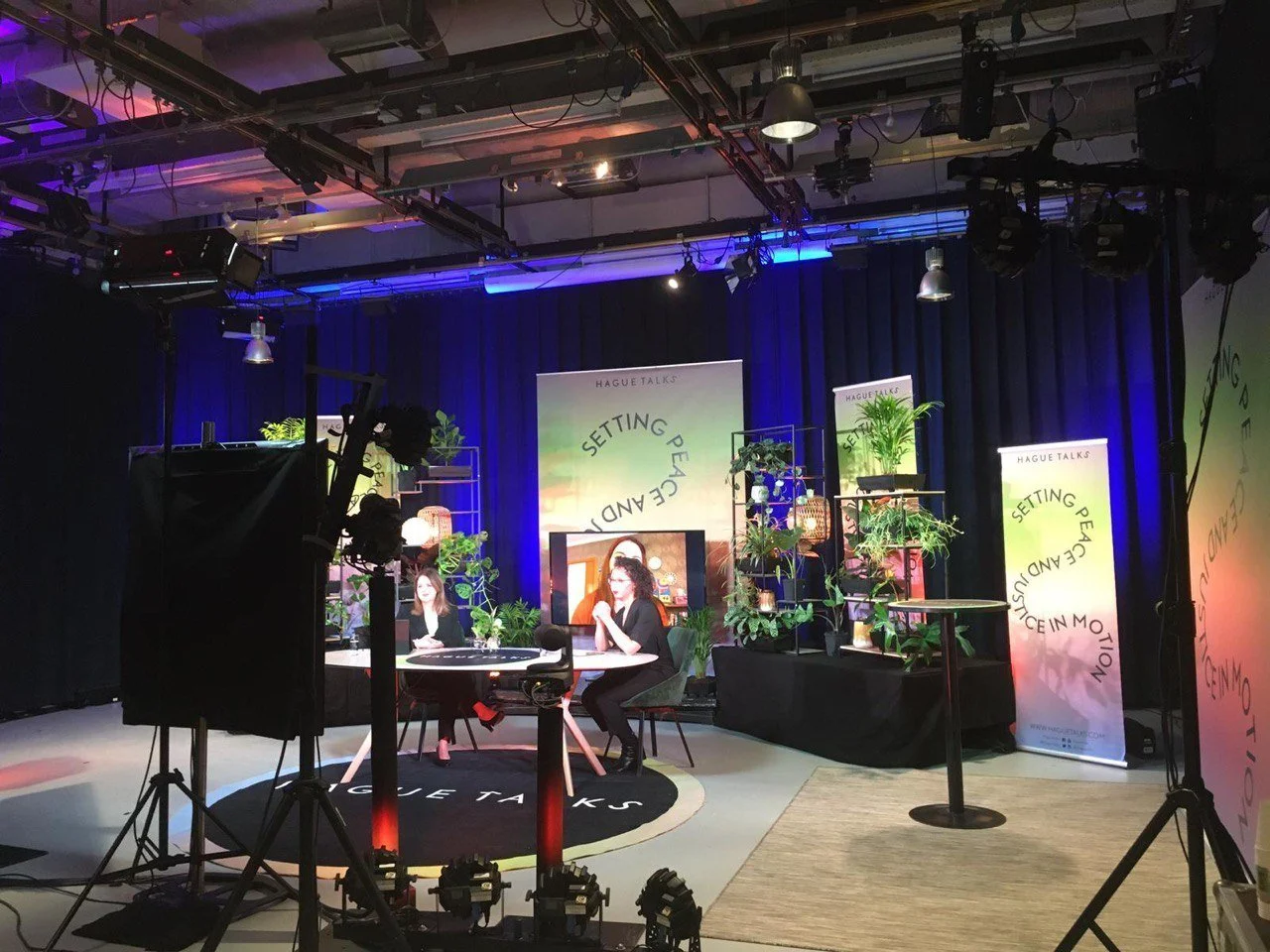 Studio voor een Hague Talks-panelgesprek met twee sprekers, planten, videofilm en kleurrijke podiumverlichting.