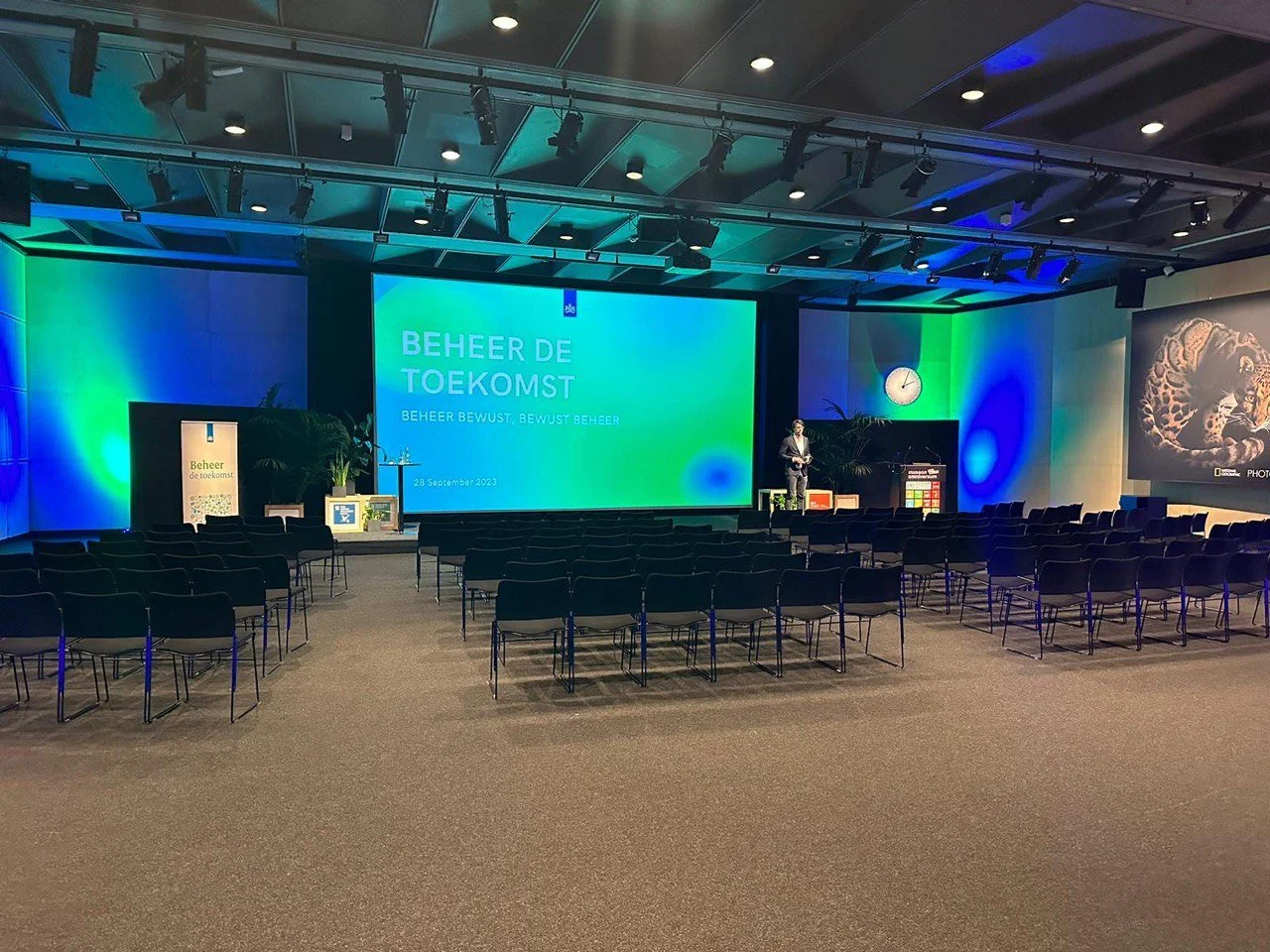 Congreszaal met podium, spreker en schermpresentatie ‘Beheer de toekomst’ tijdens een professioneel evenement.