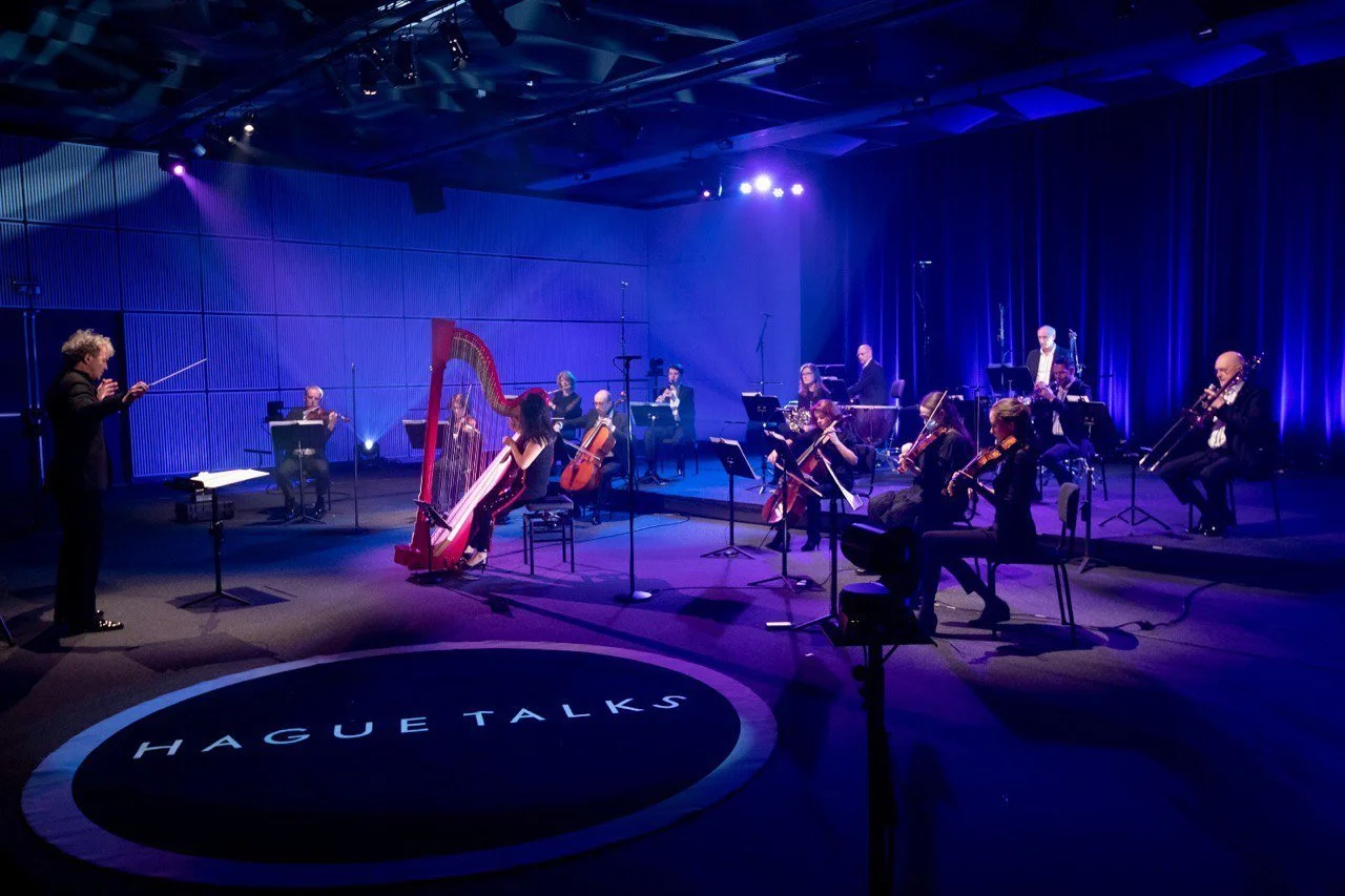 Dirigent leidt een klassiek orkest met harp, strijkers en blazers op een blauw verlicht podium van HaguelTalks.