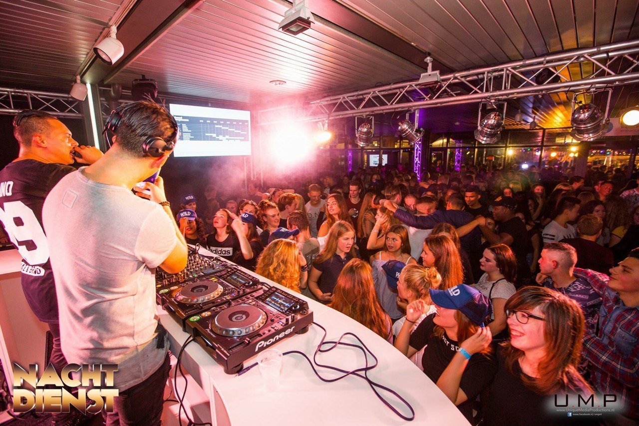 DJ draait muziek tijdens een druk nachtclubfeest met dansende menigte en kleurrijke verlichting.
