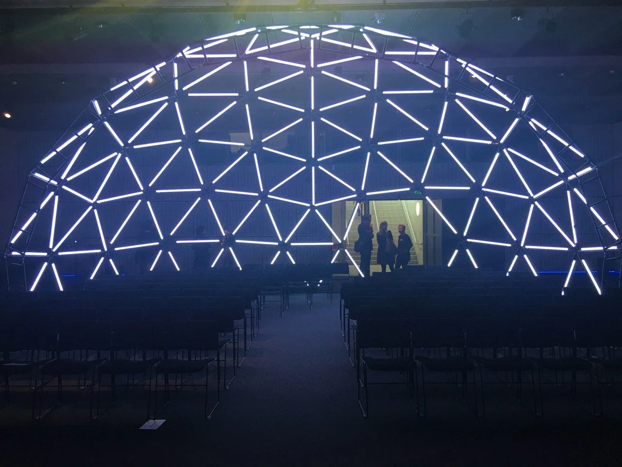 Donkere evenementenruimte met rijen stoelen en een groot halfrond podiumdecor van verlichte geometrische lijnen.