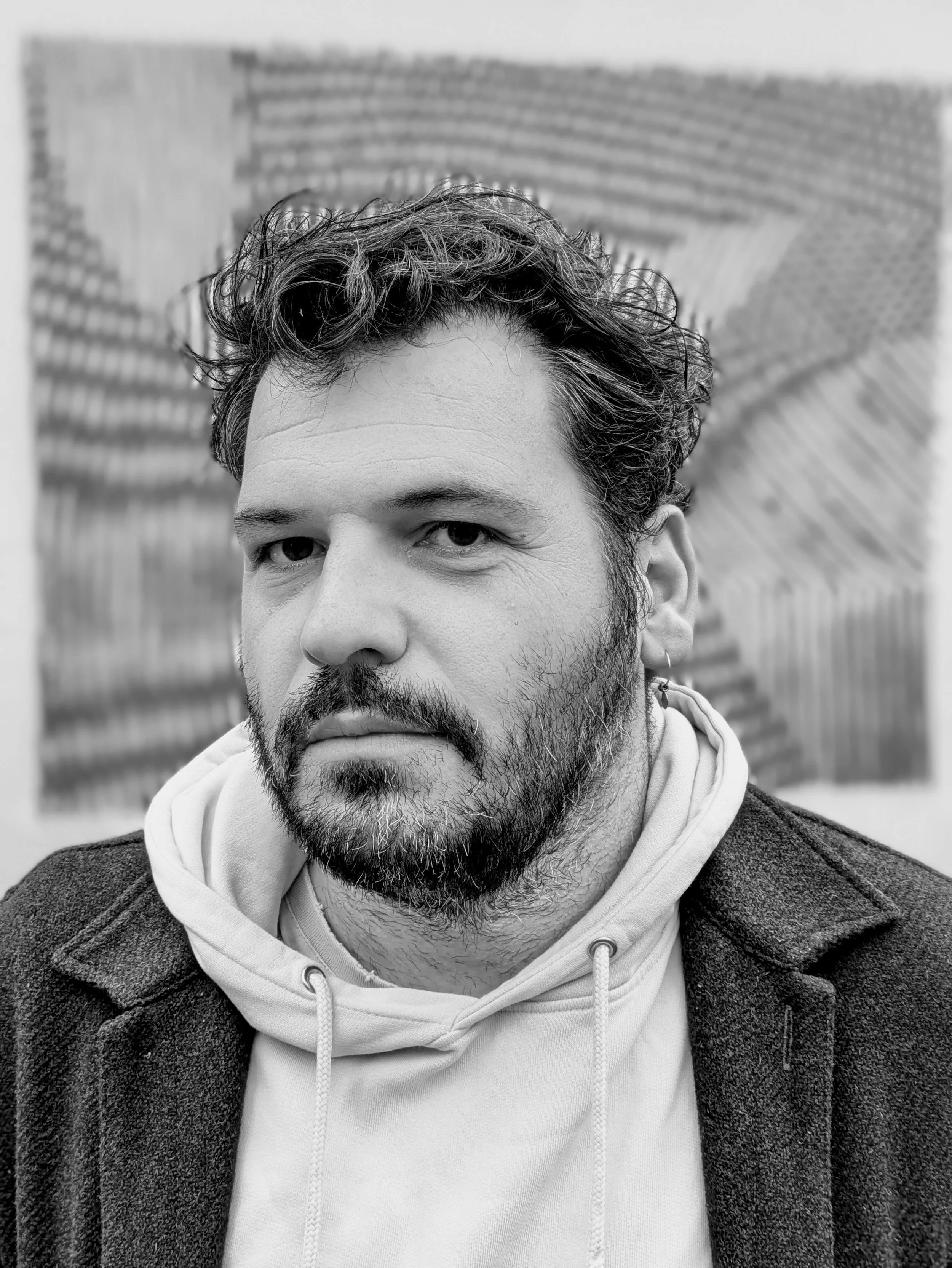 Portrait en noir et blanc en gros plan d’un homme barbu portant un sweat à capuche devant un mur de briques.