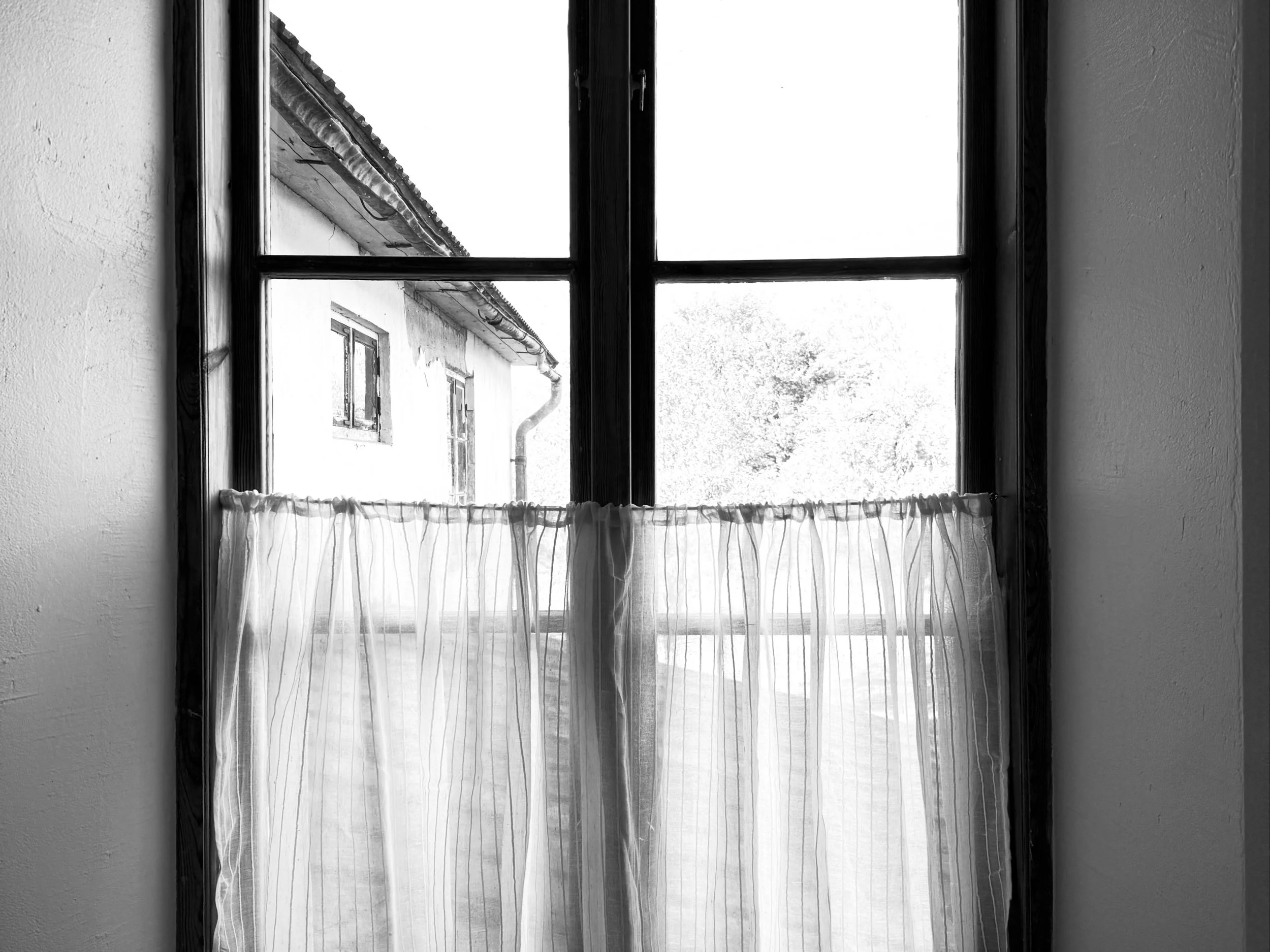 Fenêtre ancienne à croisillons avec rideau voilage, donnant sur une maison au toit de tuiles et des arbres.