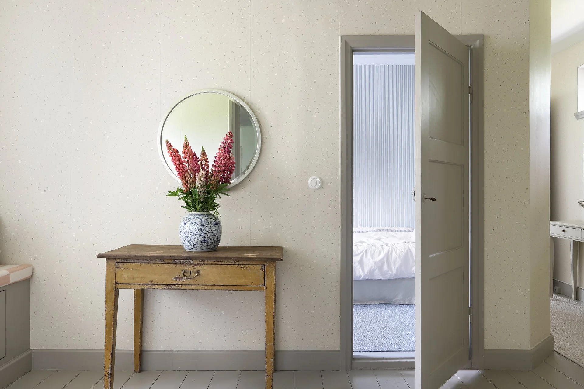 Entrée élégante avec console en bois patiné, vase fleuri et miroir rond, donnant sur une chambre.