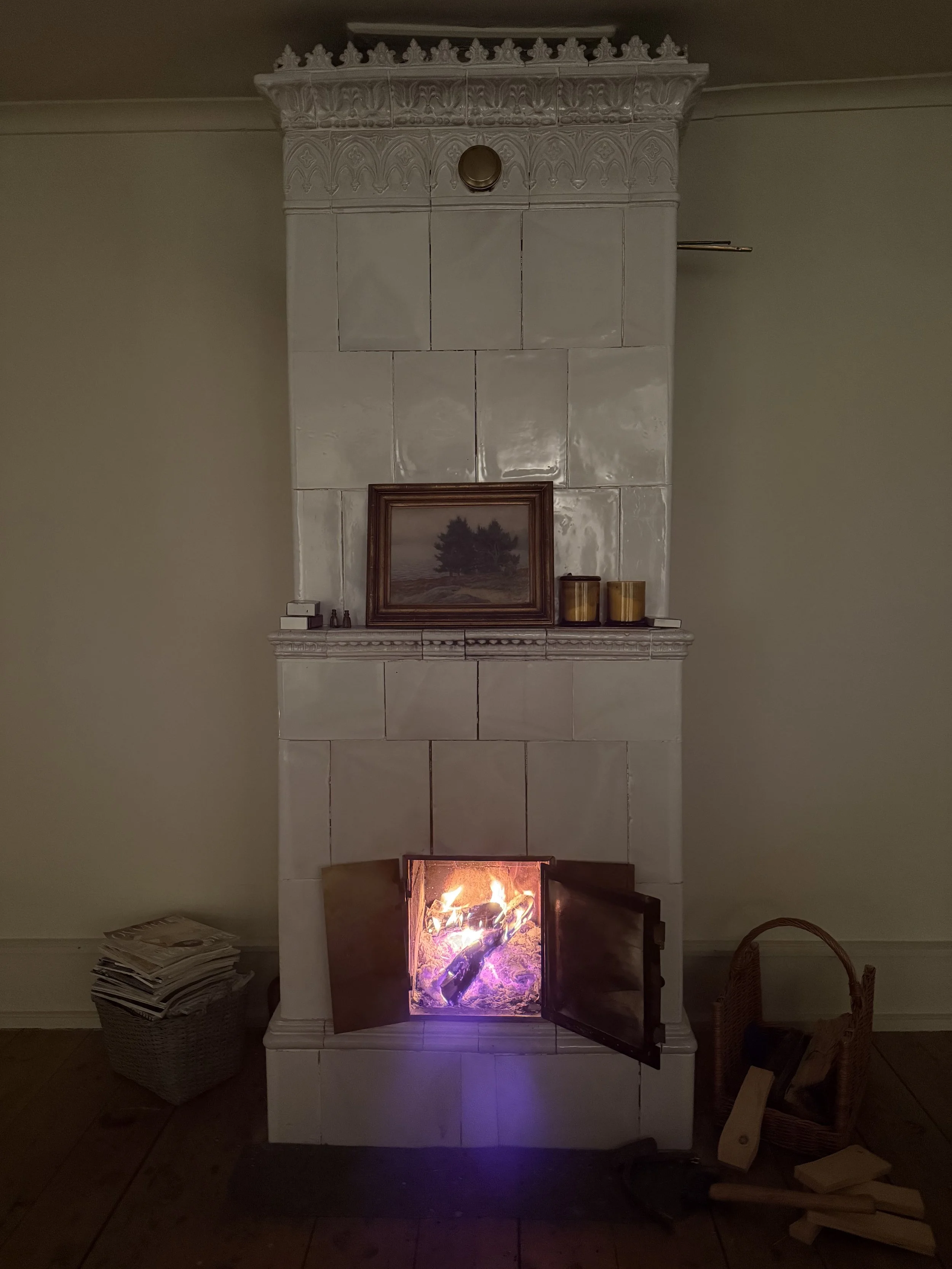 Cheminée ancienne en faïence blanche ornée, avec feu de bois allumé, tableau encadré et bougies sur le manteau.