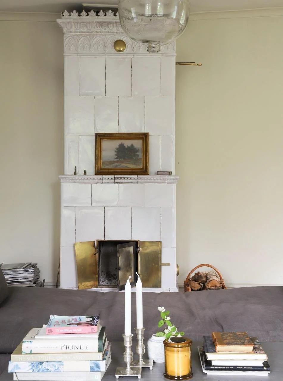 Intérieur chaleureux avec poêle en faïence blanche, tableau encadré, bougies, livres et panier de bois.