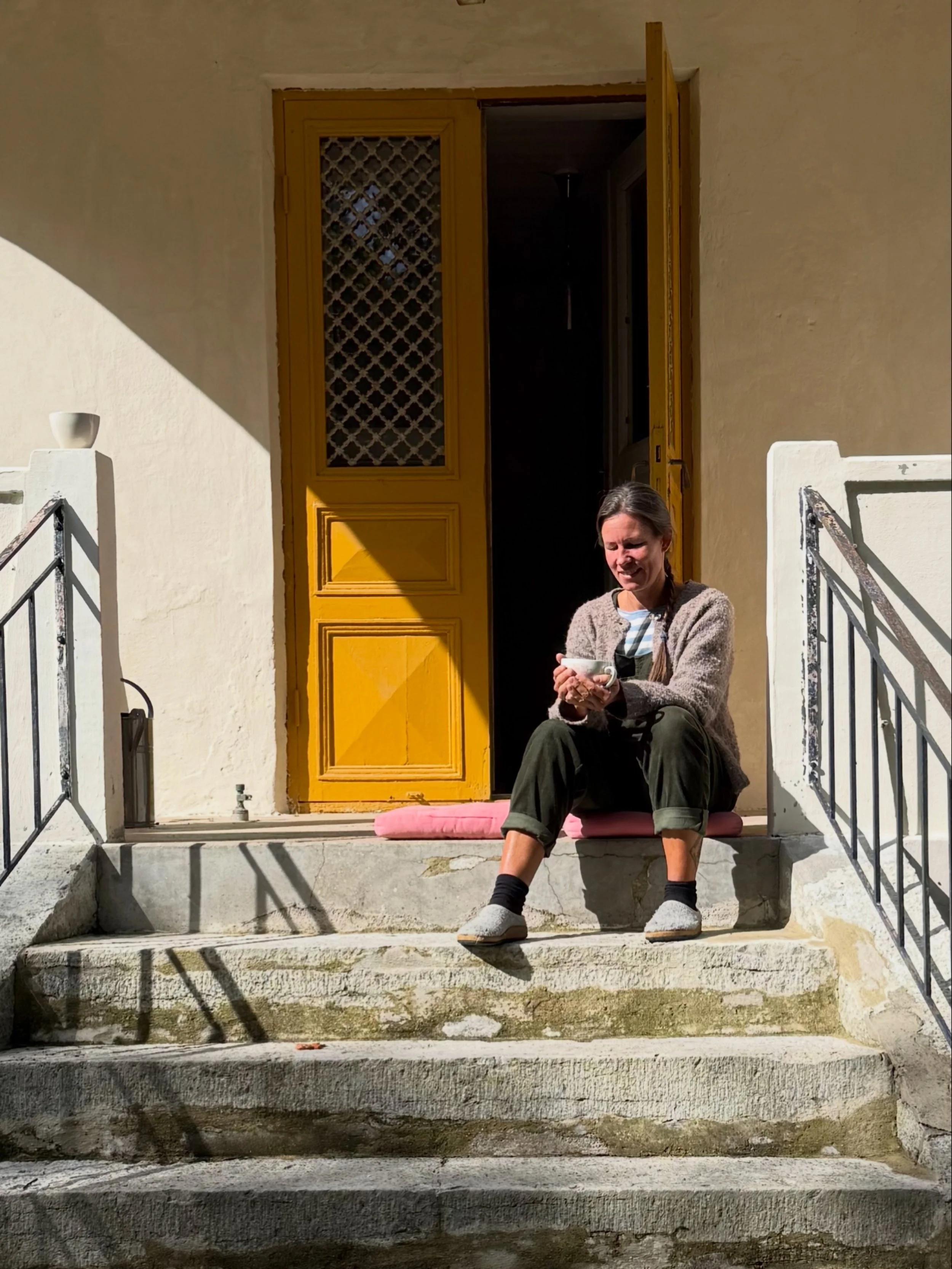 Femme assise sur les marches d’une maison, tenant une tasse devant une porte jaune ouverte.