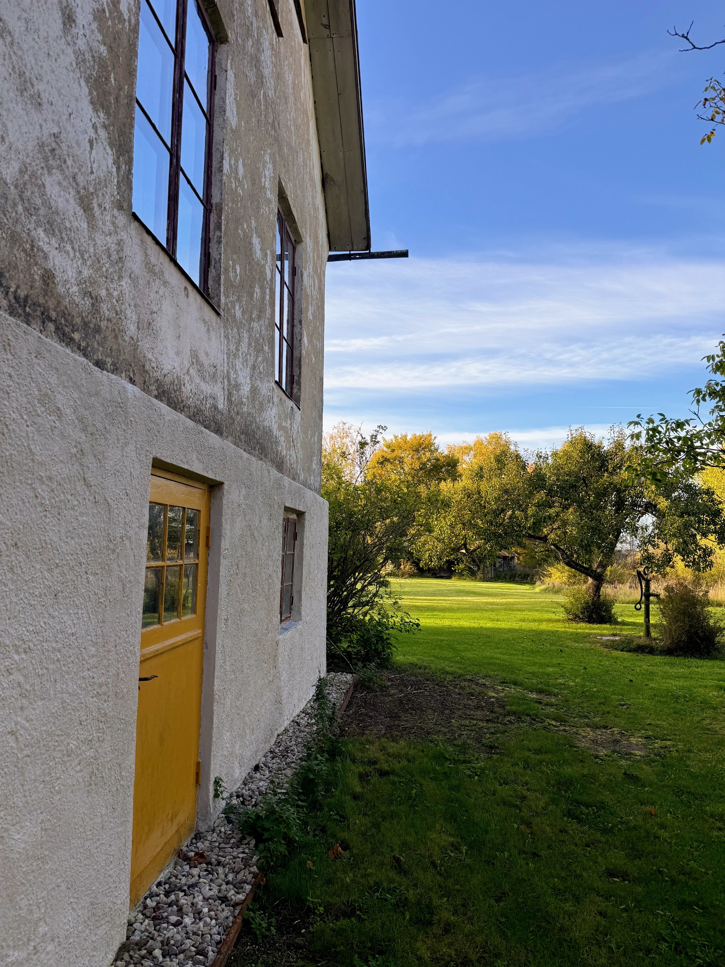 Ancienne maison aux murs blanchis et à la porte jaune, donnant sur un jardin verdoyant sous un ciel bleu.