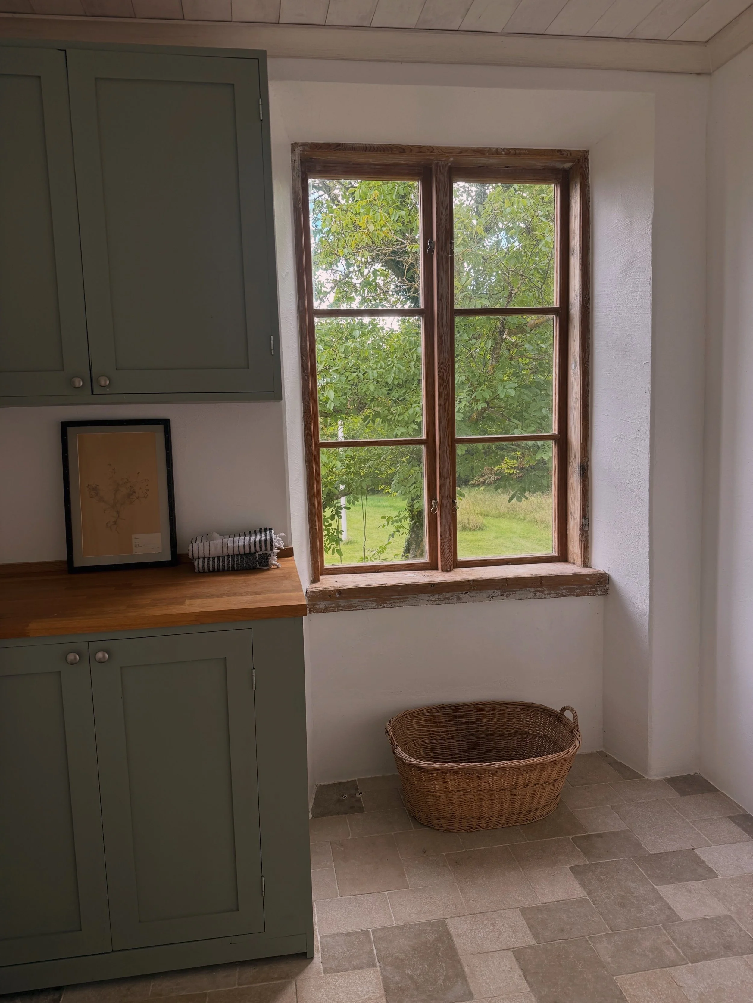 Intérieur rustique avec fenêtre en bois donnant sur un jardin, placards vert olive, œuvre encadrée et panier en osier.