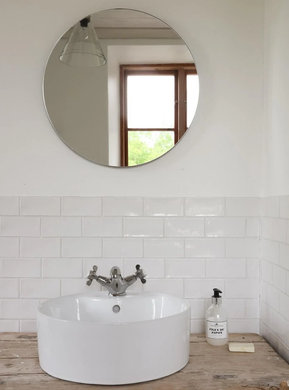 Salle de bains avec lavabo blanc, robinet chromé, miroir rond et carrelage mural blanc.
