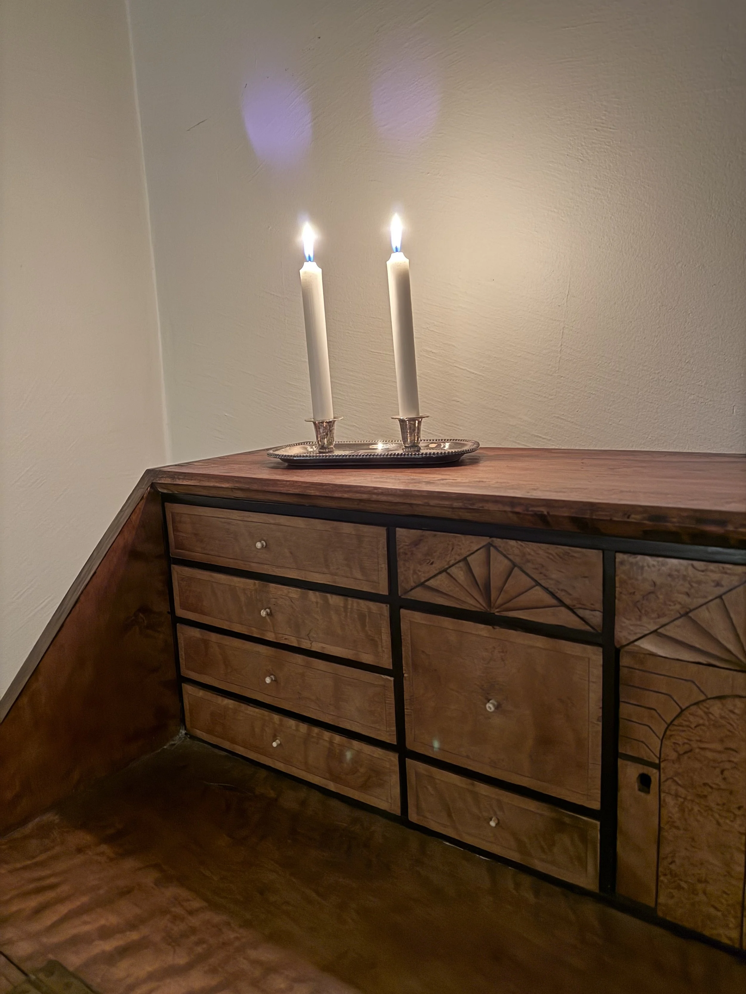 Deux bougies blanches allumées sur un plateau argenté posé sur une commode en bois.