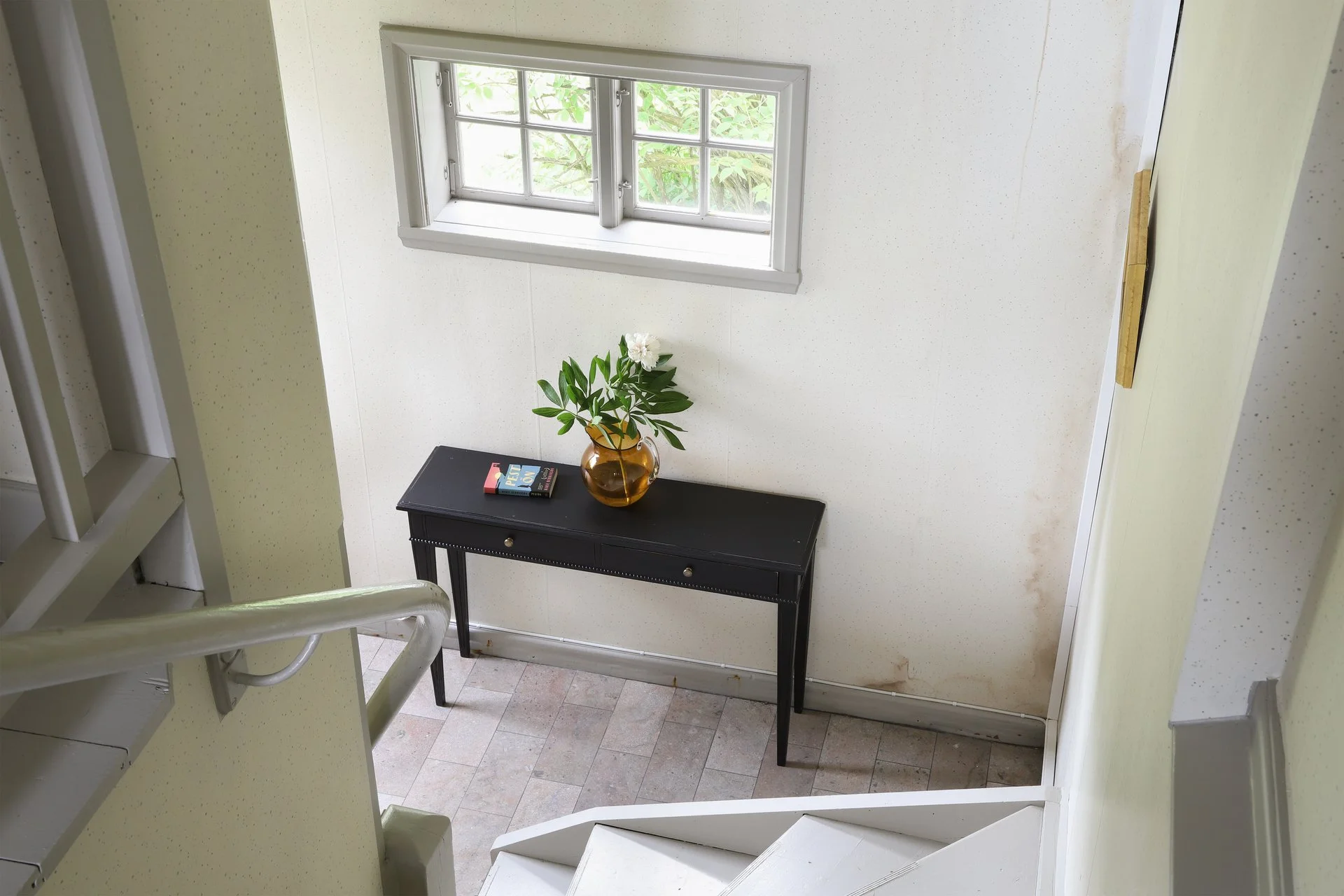 Palier d’escalier avec console noire, vase de fleurs blanches et fenêtre à petits carreaux.