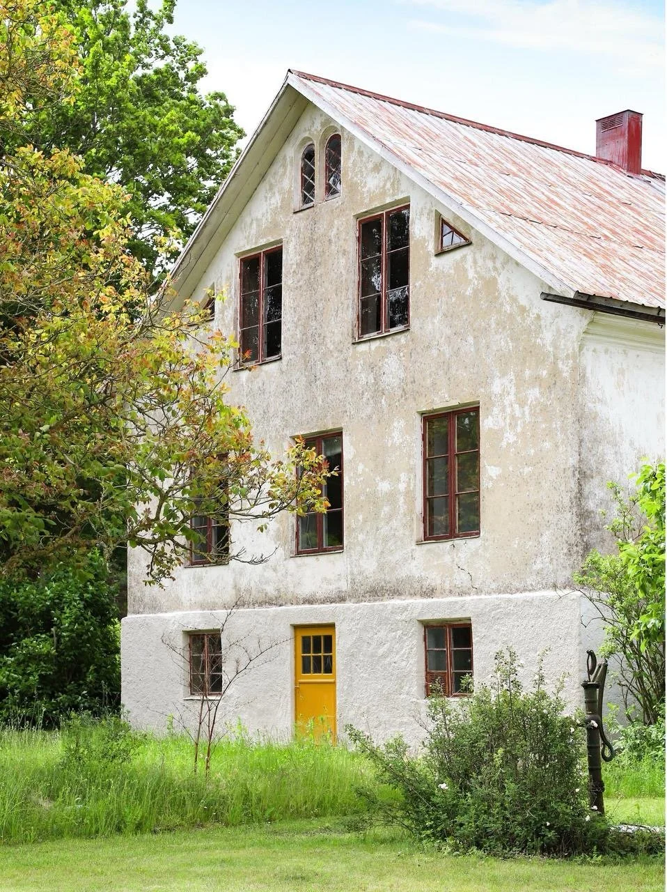 Ancienne maison blanche décrépie avec toit en tôle rouillée, porte jaune et fenêtres rouges, entourée d’une végétation luxuriante.