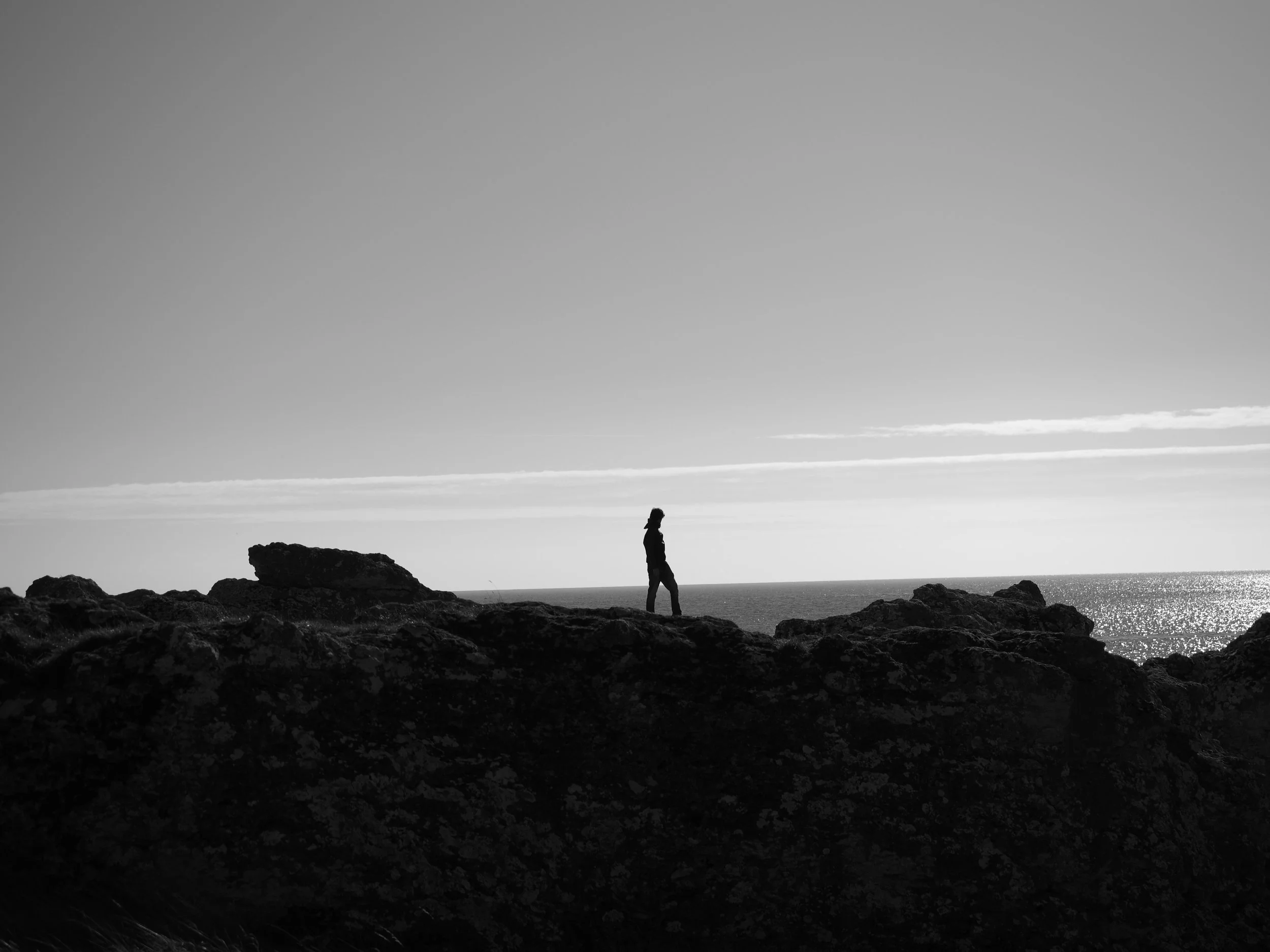 Silhouette d’une personne marchant sur des rochers au bord de la mer, sous un ciel nuageux, en noir et blanc.