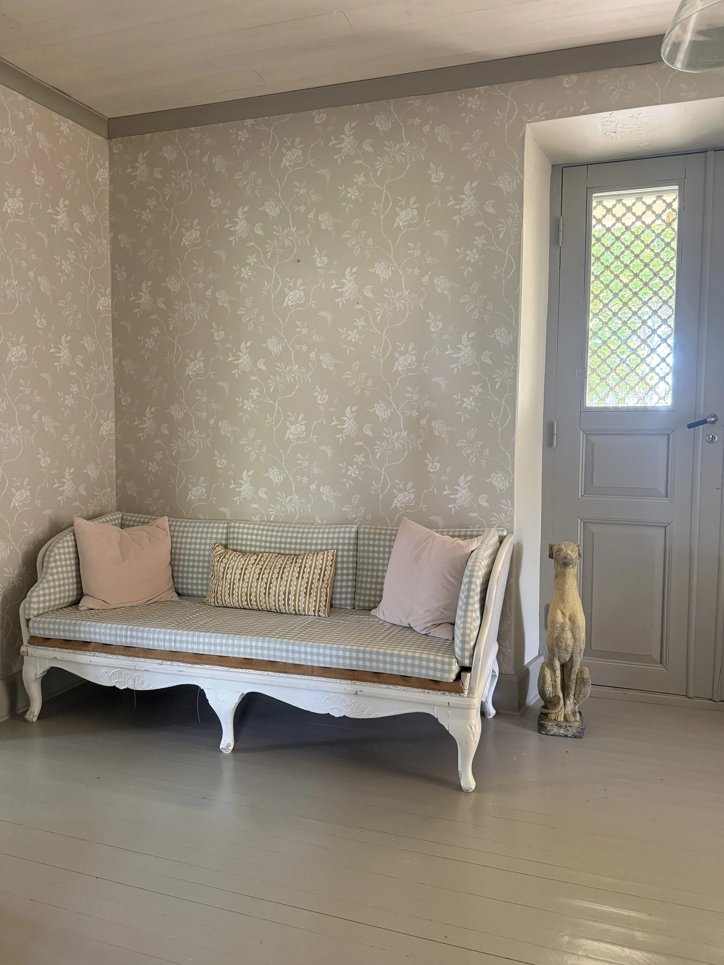 Salon élégant avec canapé vintage blanc à coussins, papier peint floral et porte bleue vitrée.