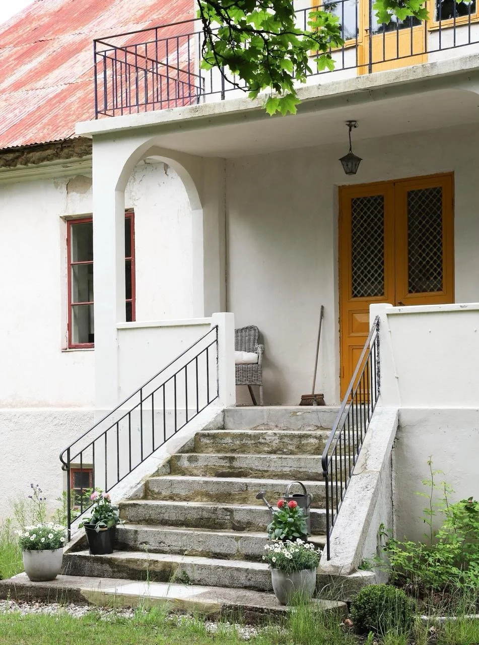 Entrée d’une maison ancienne avec escalier en pierre, balustrades noires, porte en bois et jardinières fleuries